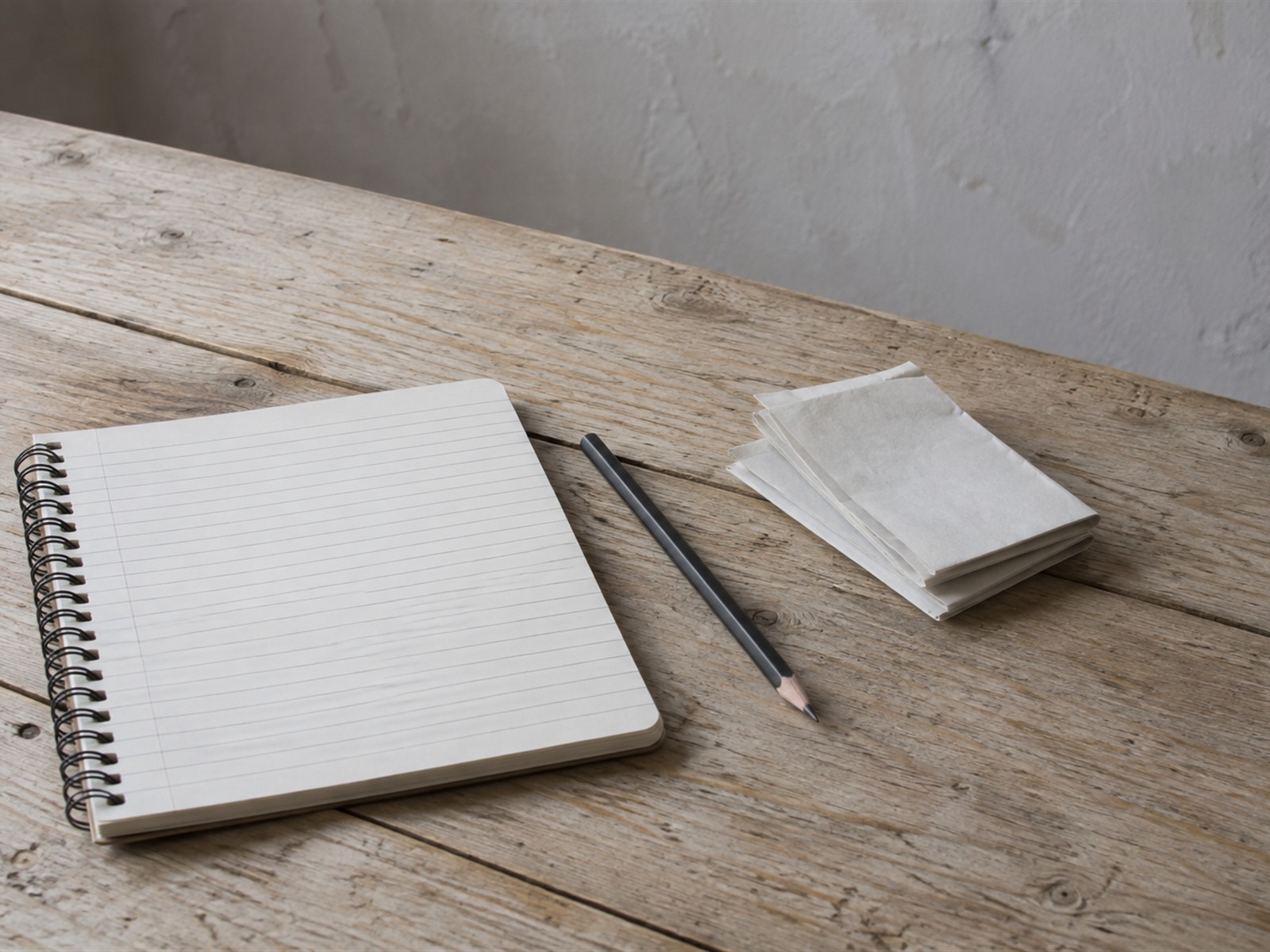 A blank ruled notebook, a sharpened pencil and a small stack of folded paper receipts on a pale wooden table.