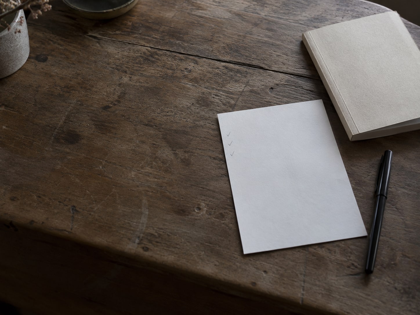 A sheet of blank paper with faint pencil check marks in the margin, beside a closed notebook and a pen on a wooden desk.