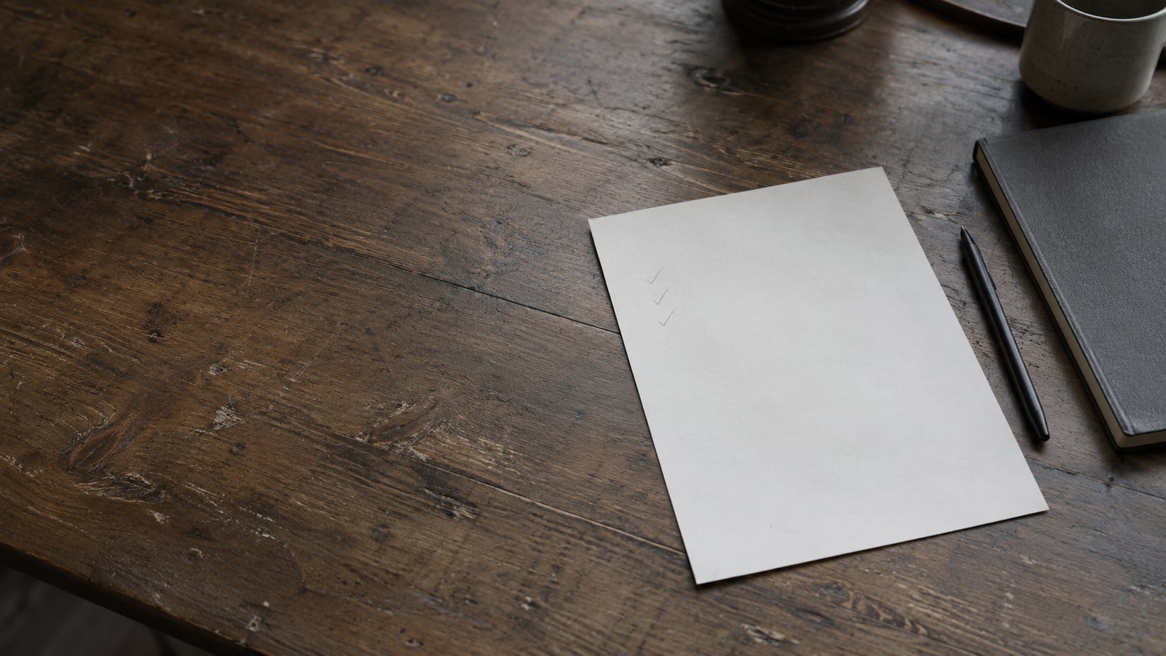 A blank sheet of paper with faint pencil check marks beside a closed notebook and a pen on a wooden desk.