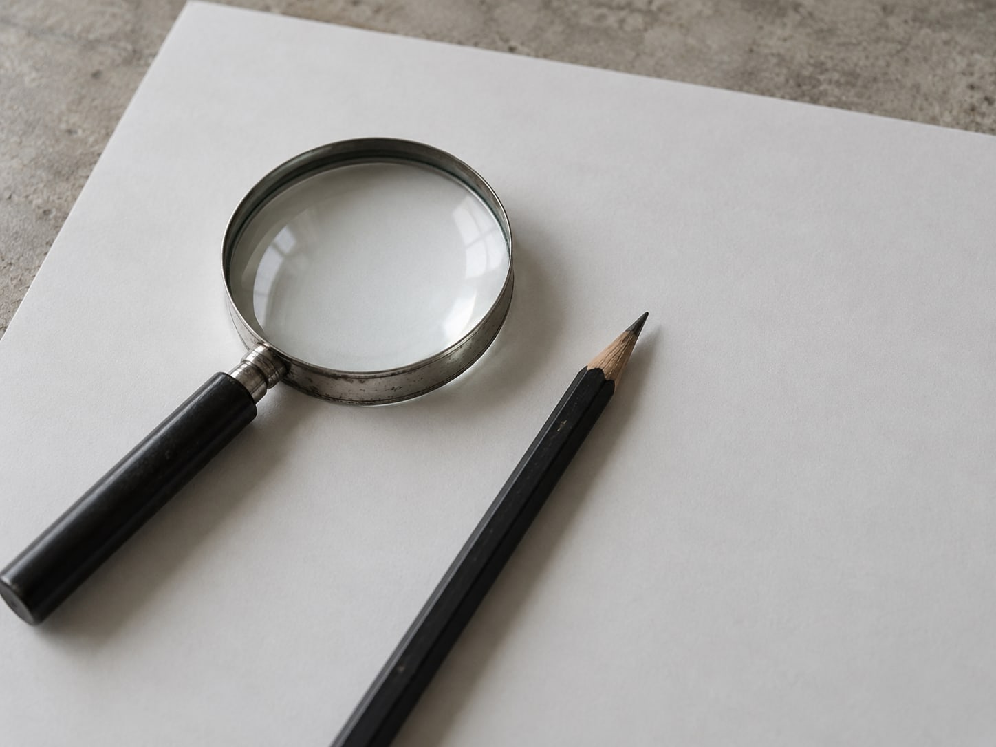 A magnifying glass resting on a blank sheet of paper beside a sharpened pencil.