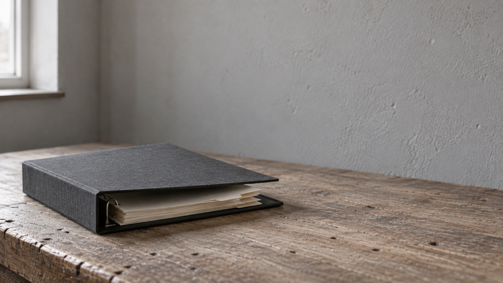 A dark ring binder of papers lying on a worn wooden desk by a window.