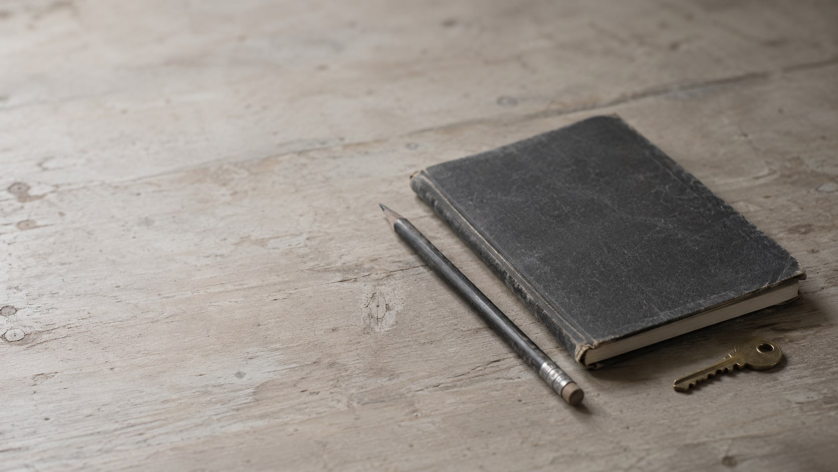 A closed notebook, a pencil and a small brass key resting on a clean wooden counter in soft window light.