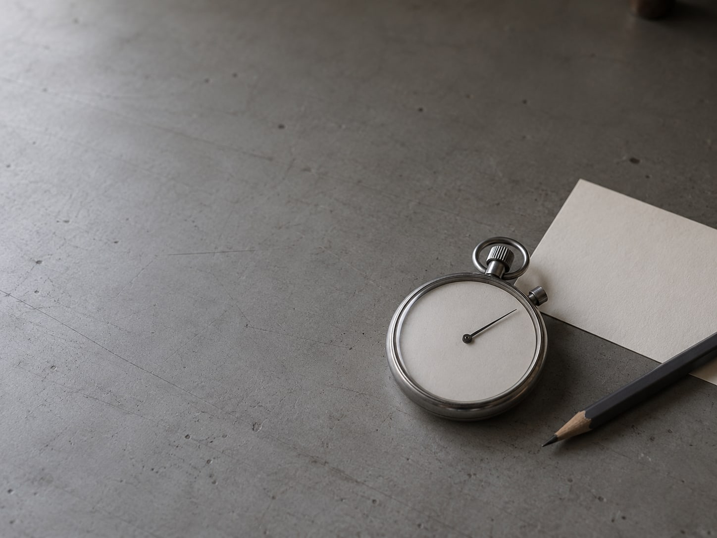 A mechanical stopwatch lying beside a pencil and a blank index card on a gray desk.