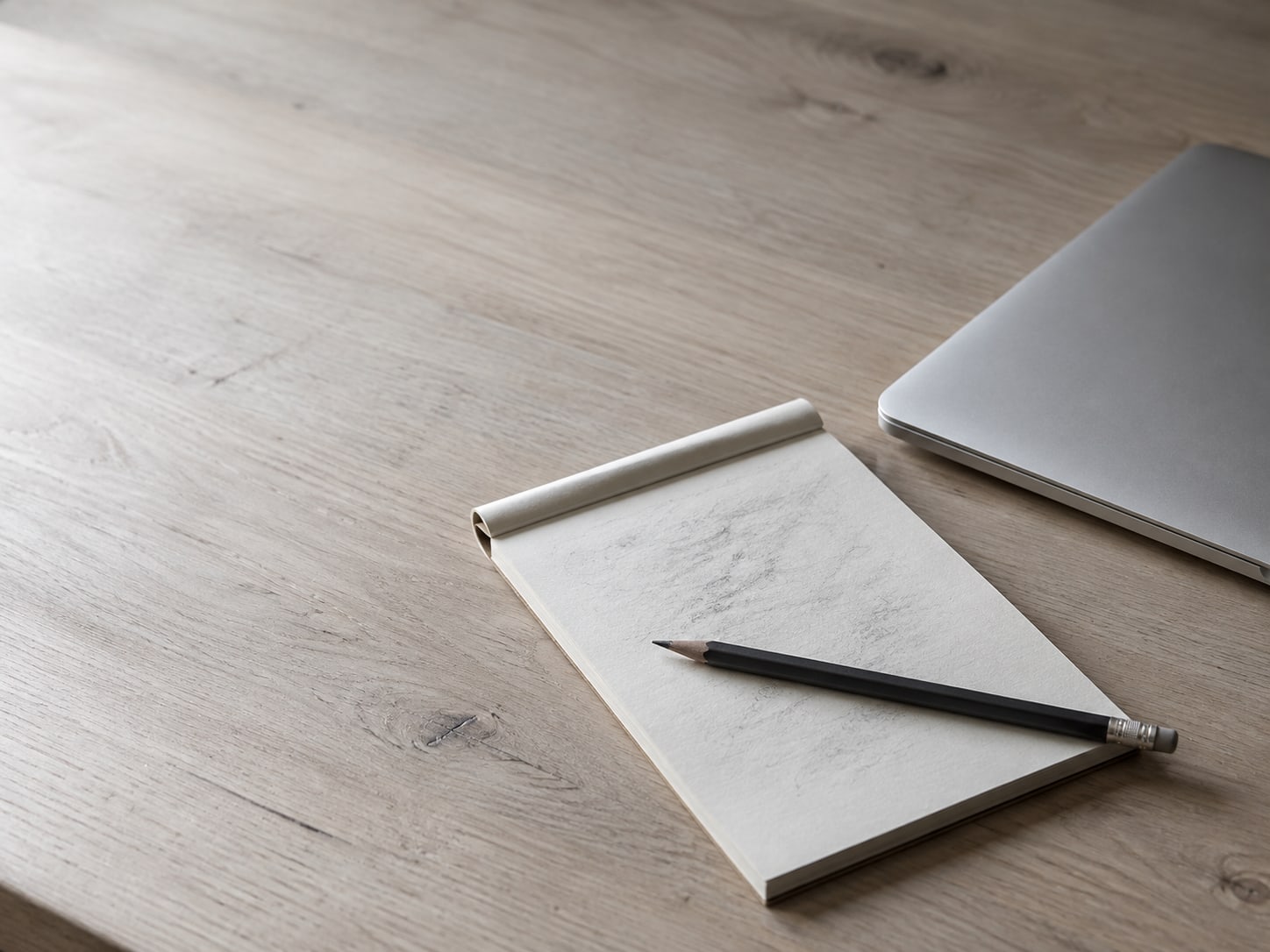 Una laptop cerrada junto a un cuaderno cubierto de marcas de lápiz tenues e ilegibles y un lápiz atravesado sobre la página, en un escritorio de madera.