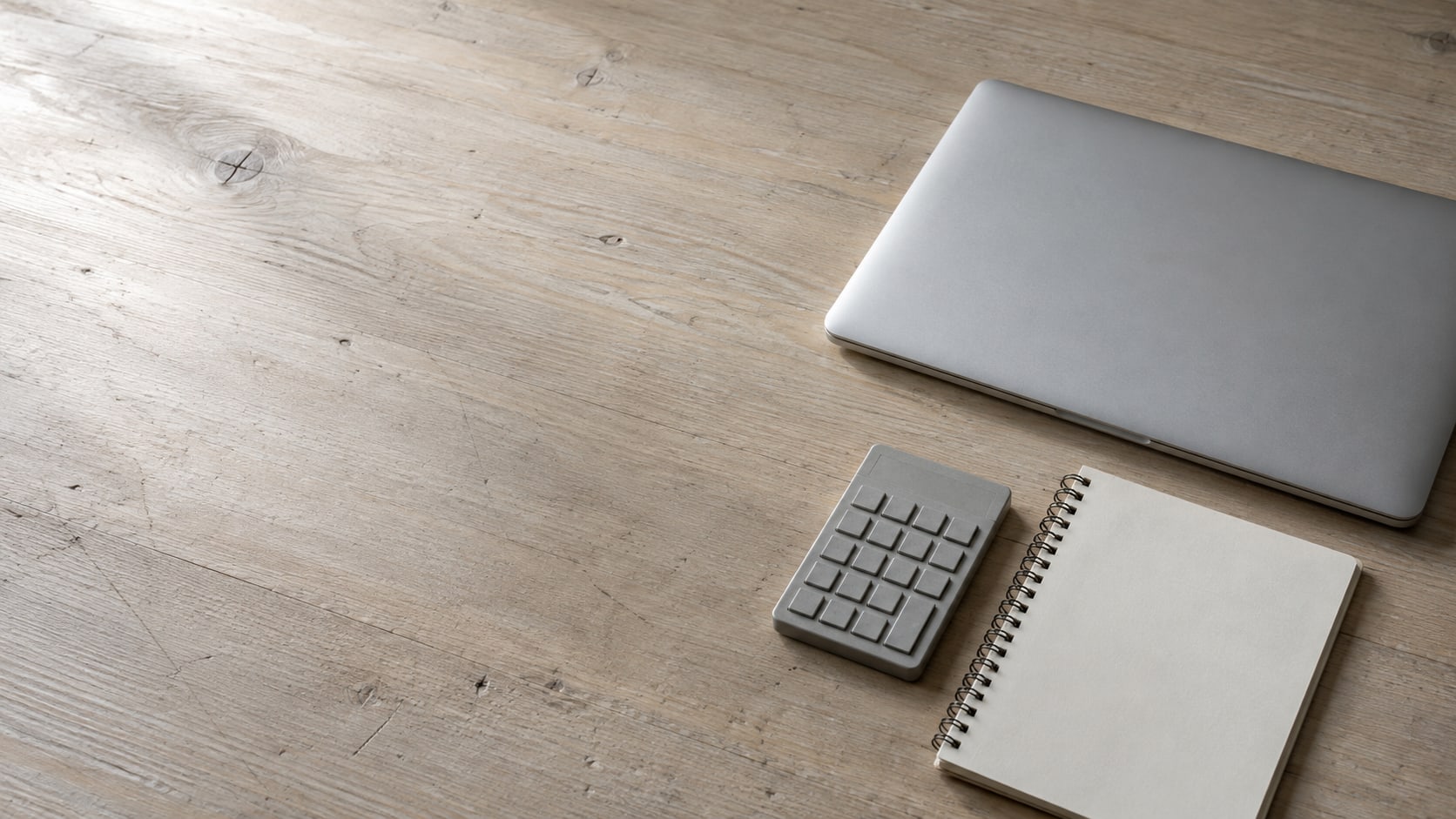 Una laptop cerrada, una pequeña calculadora y un cuaderno en blanco acomodados sobre un escritorio de madera con luz suave.