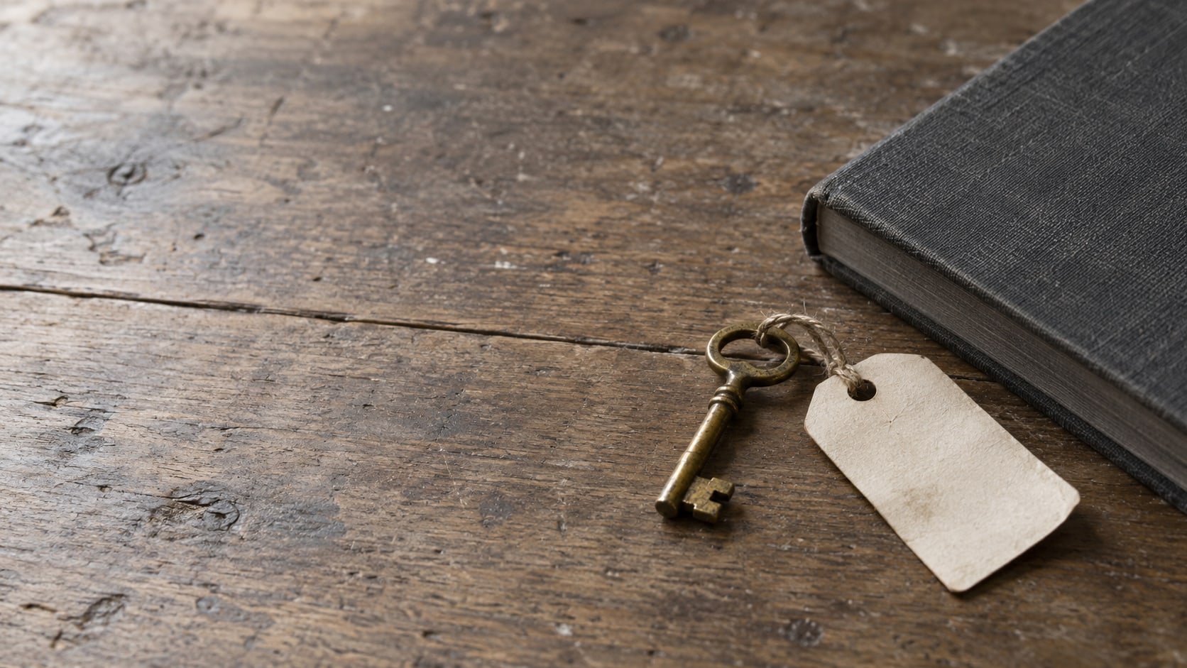 A small brass key and a blank paper tag resting on a worn wooden desk beside a closed notebook.