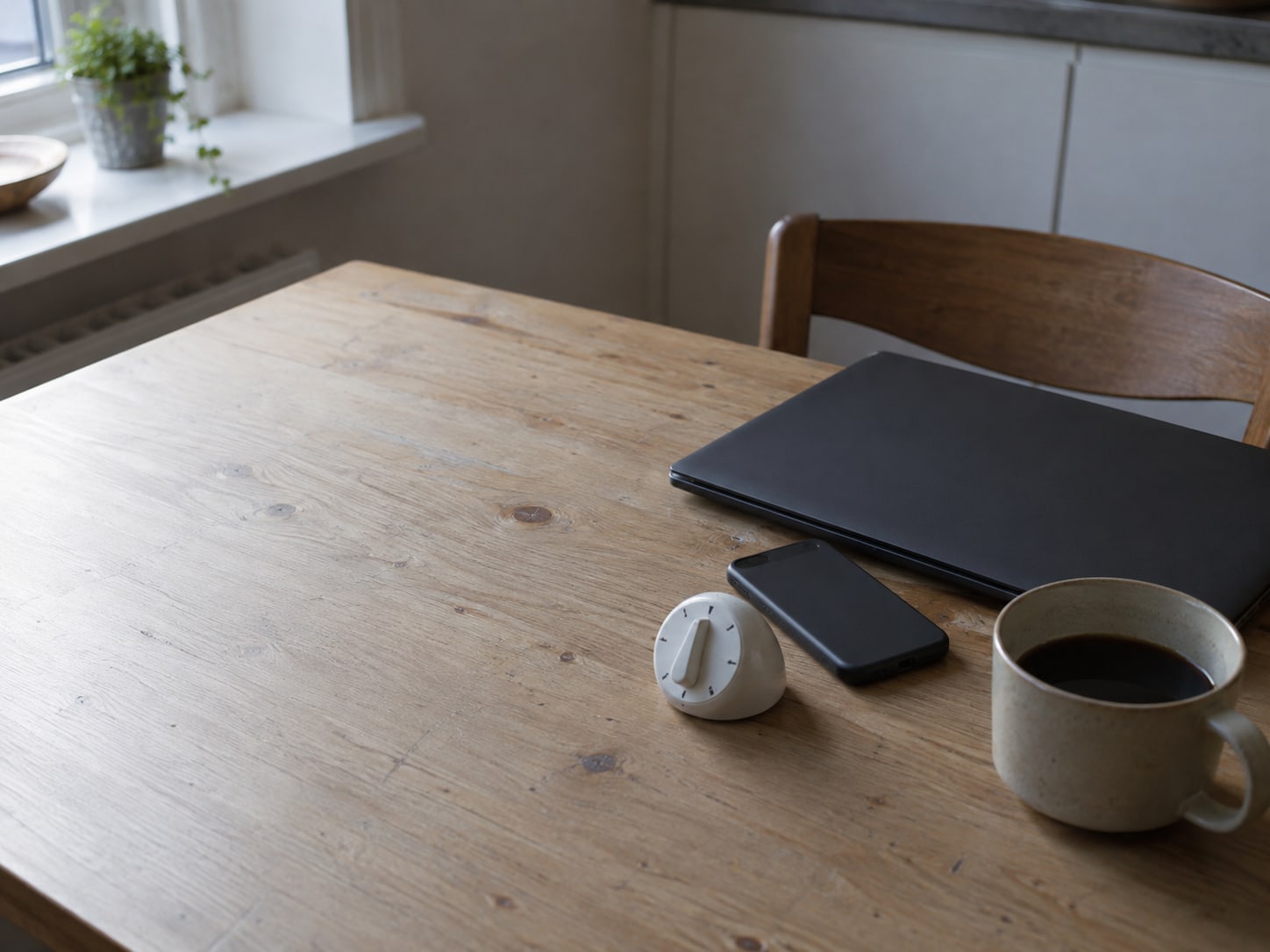 A closed laptop and a phone lying face down on a kitchen table beside a simple white kitchen timer and a mug of coffee.