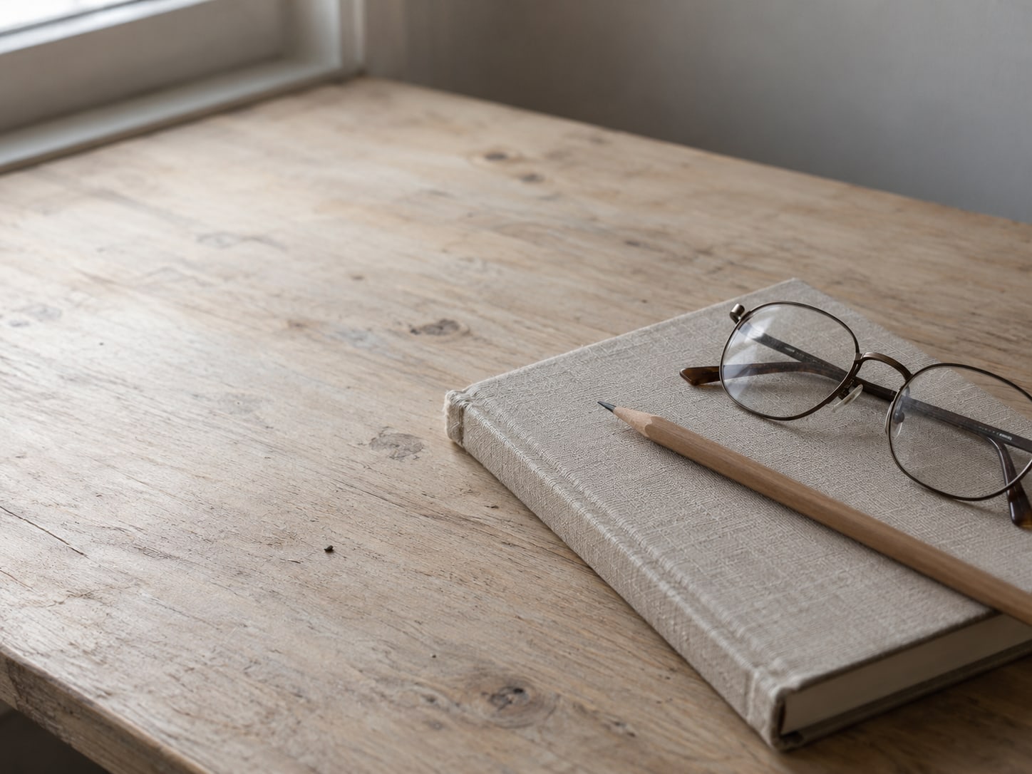 A closed hardcover notebook with a pencil and a pair of reading glasses on a clean desk.