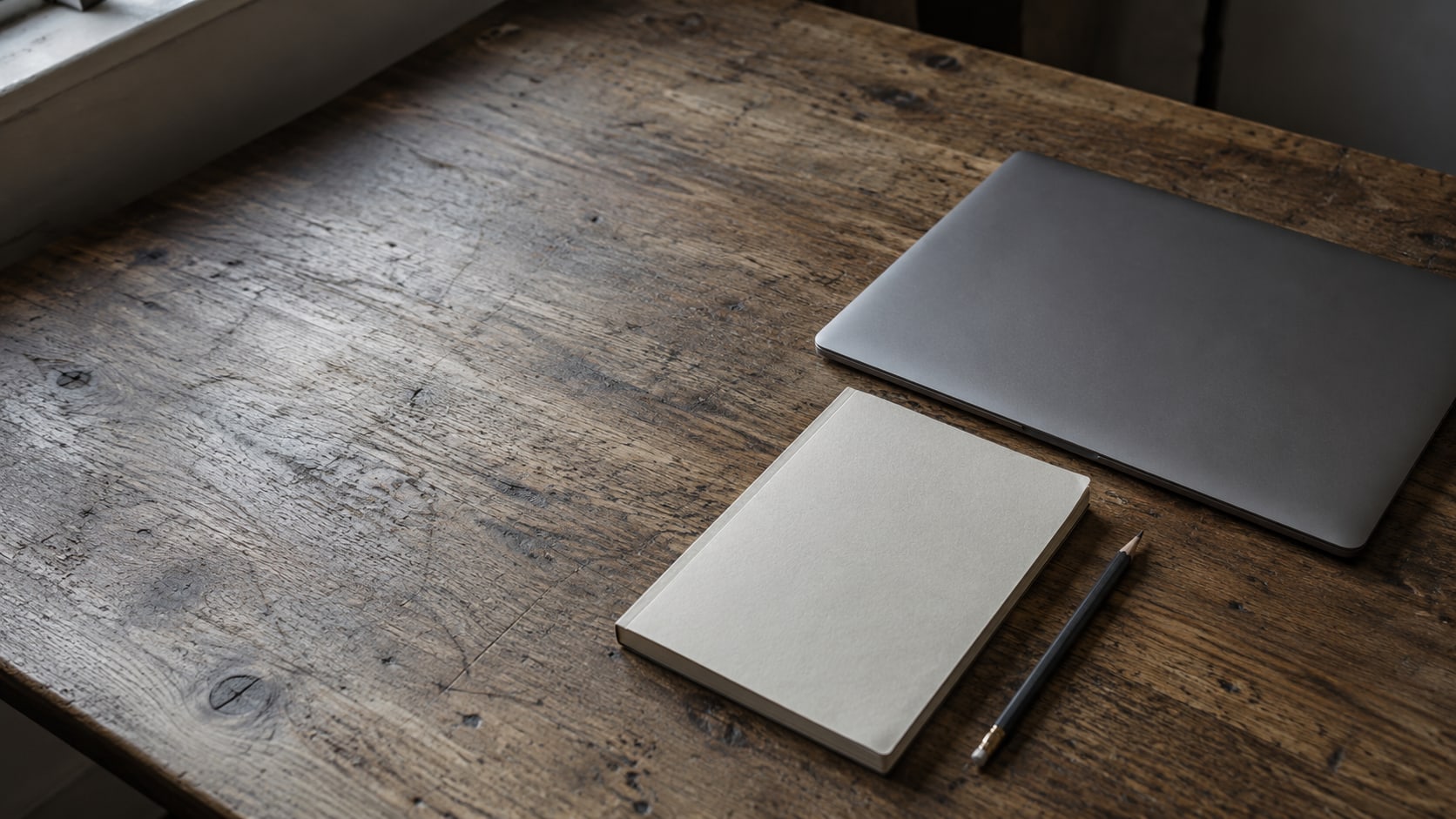 A closed laptop beside a blank notebook and a pencil on a worn wooden desk.