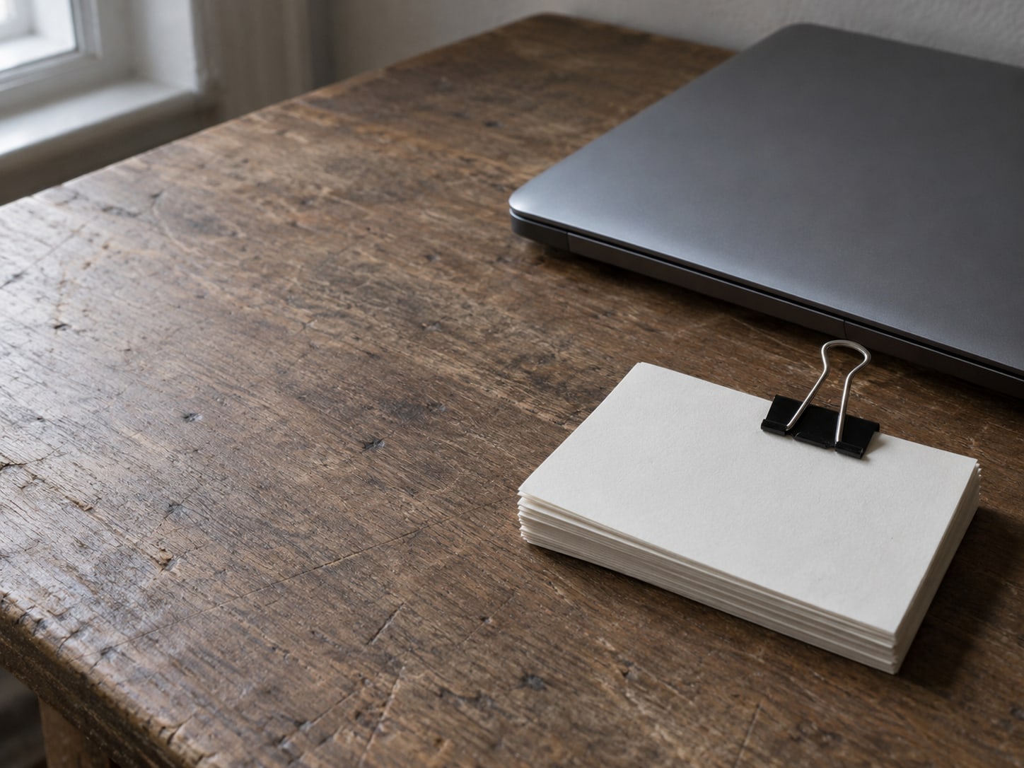 A small stack of unmarked index cards held by a binder clip next to a closed laptop on a desk.