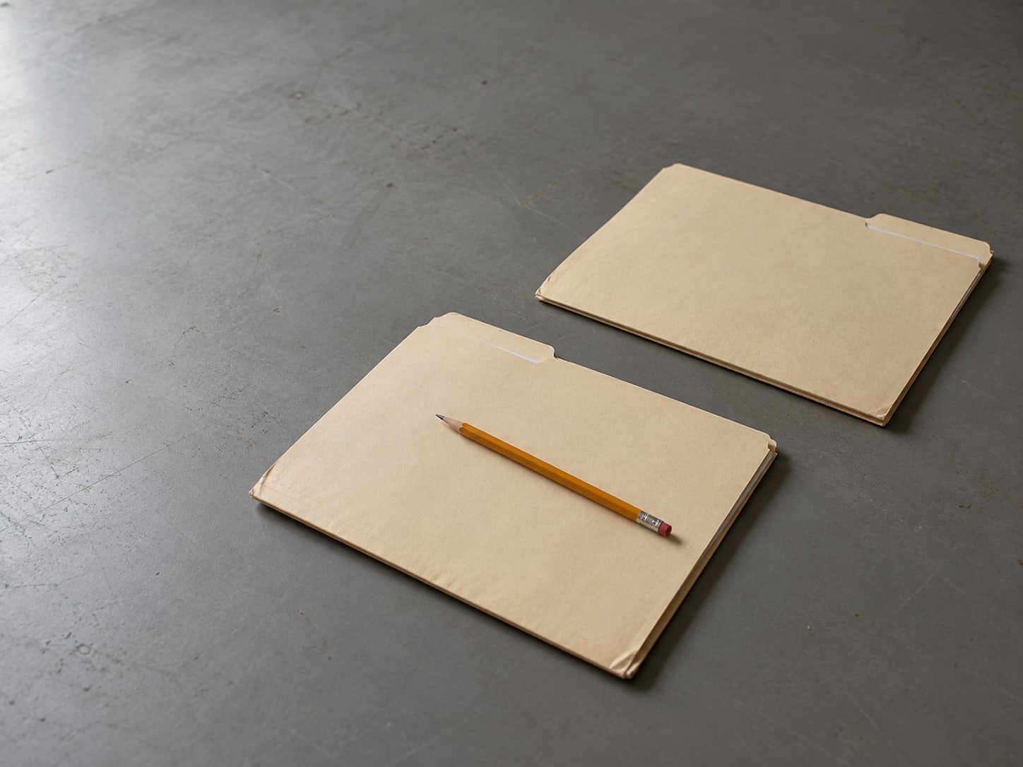 Two closed manila folders on a gray table, one pushed a few inches apart from the other, with a sharpened pencil on top of the nearer one.