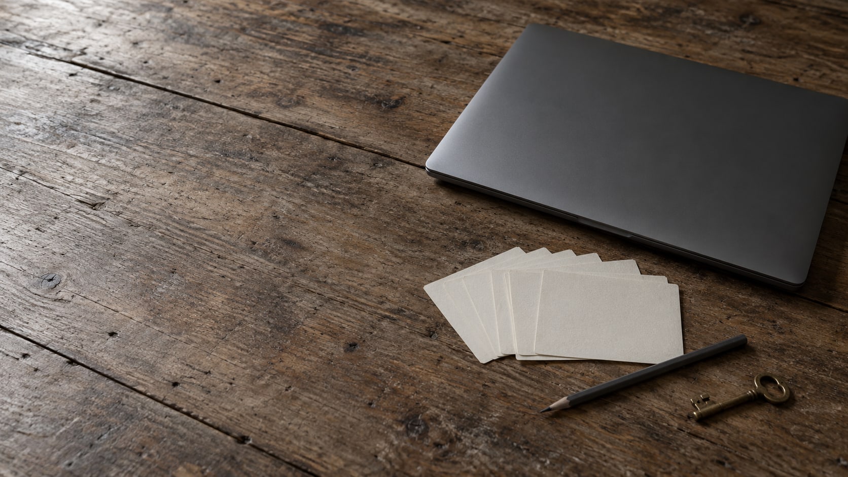 A closed laptop on a wooden desk with a fanned row of blank index cards, a pencil and a brass key beside it.