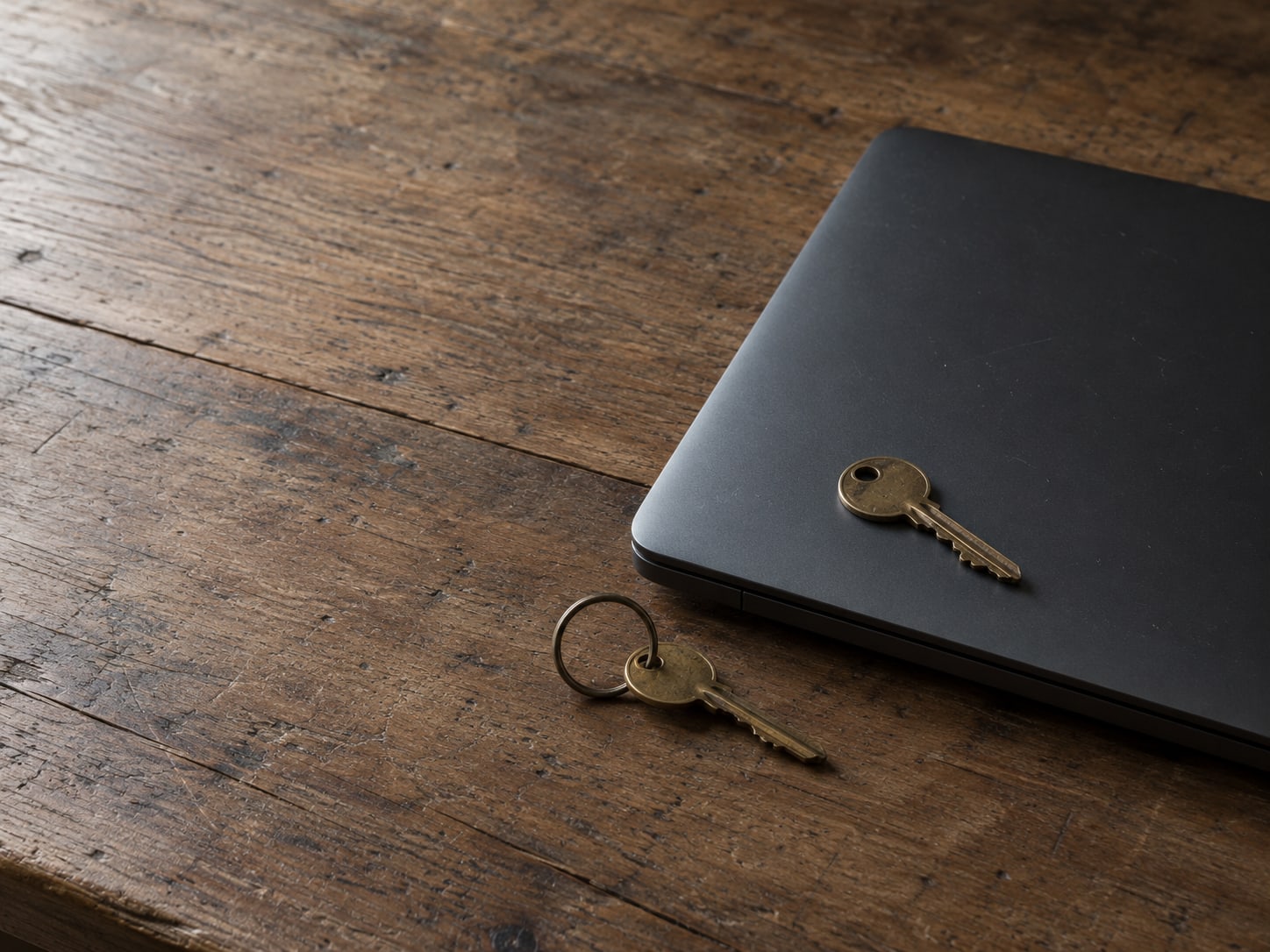 A single brass key resting on a closed laptop on a worn wooden desk, with a second key on a ring beside it.
