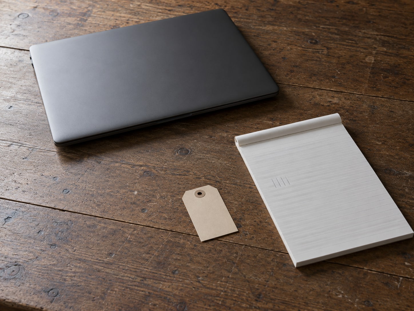 A closed laptop and a small paper tag on a wooden desk, with a ruled notepad holding faint pencil tick marks.