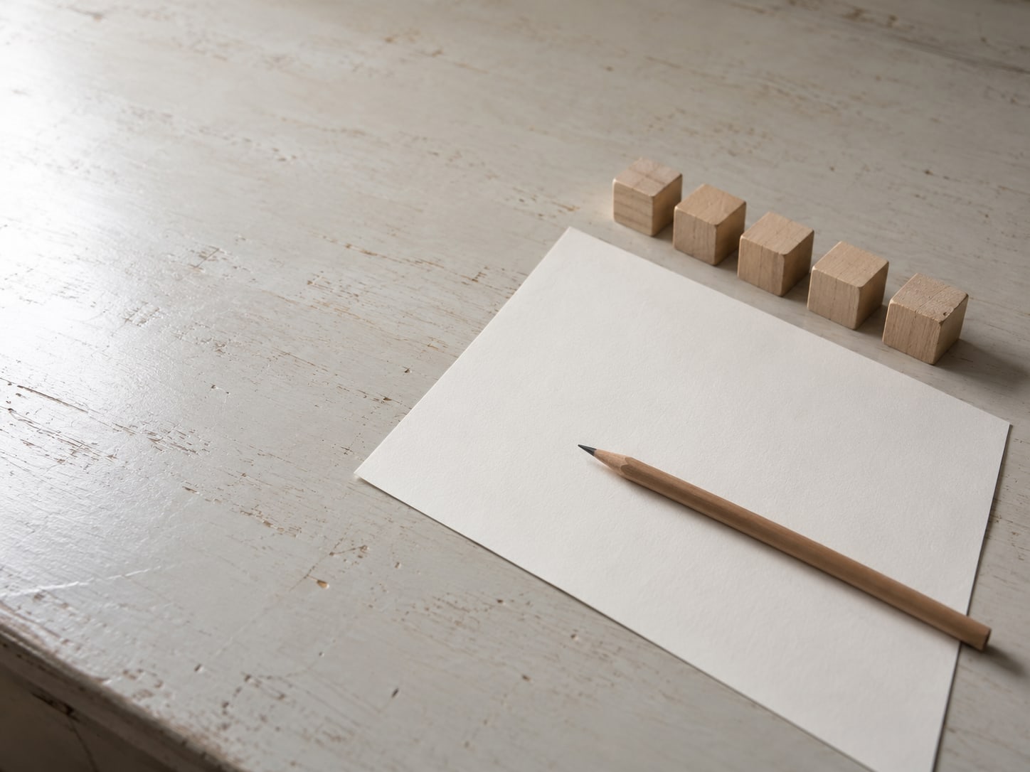 A single sheet of blank paper with a pencil resting across it beside five small, evenly spaced wooden blocks.