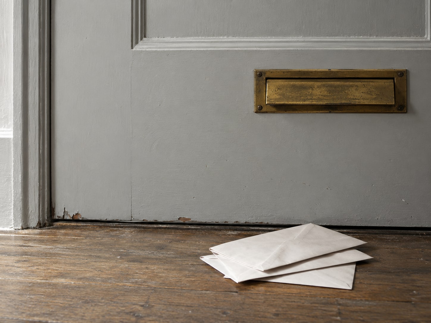 A brass mail slot set into a plain painted door, with a few blank envelopes resting on the floor below it.