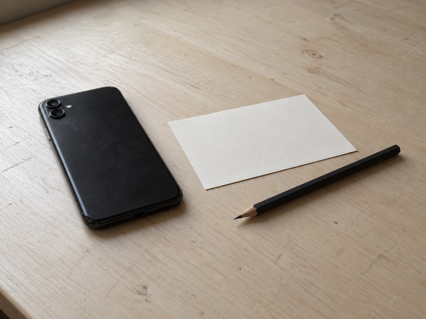 A closed phone lying beside a blank index card and a sharpened pencil on a pale desk.
