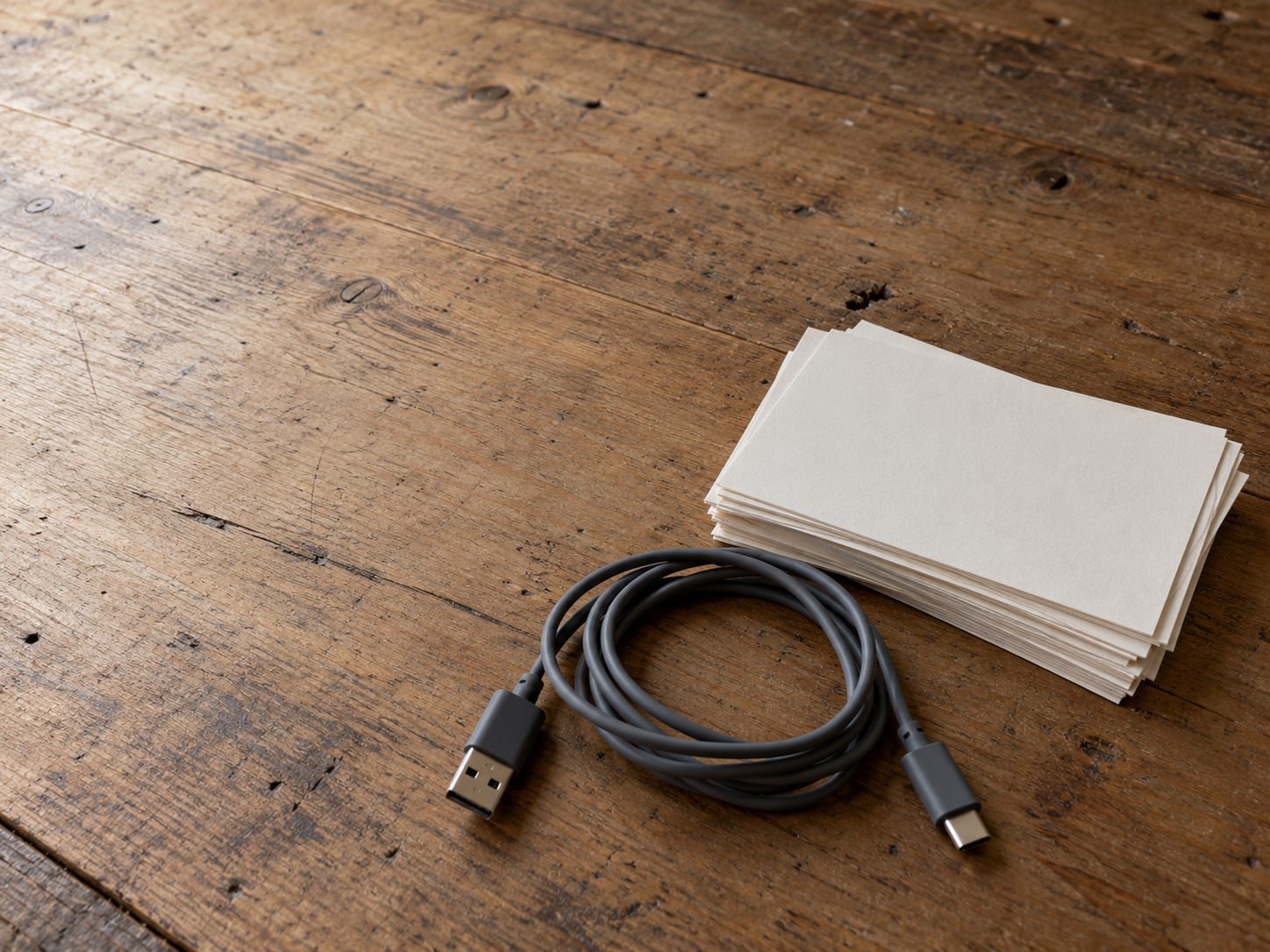 A phone charger lying coiled beside a small stack of blank index cards on a work table.