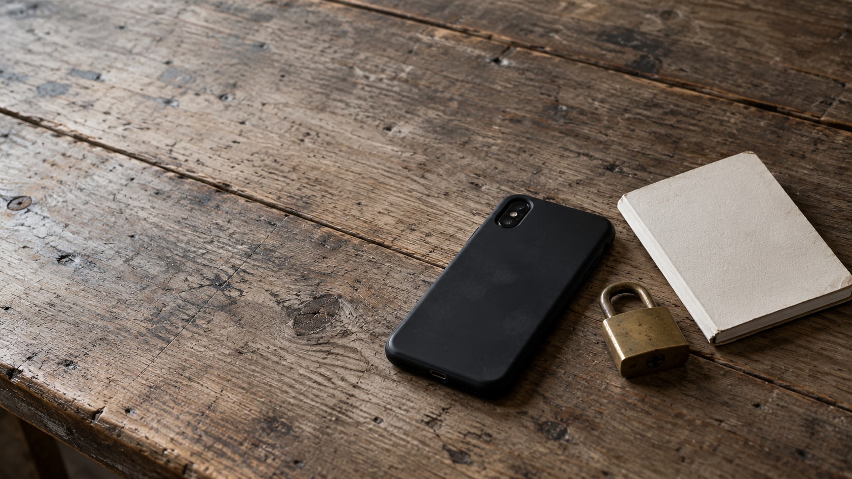 Un teléfono cerrado boca abajo sobre un mostrador de madera de una tienda, junto a un candado de latón y una pequeña libreta.