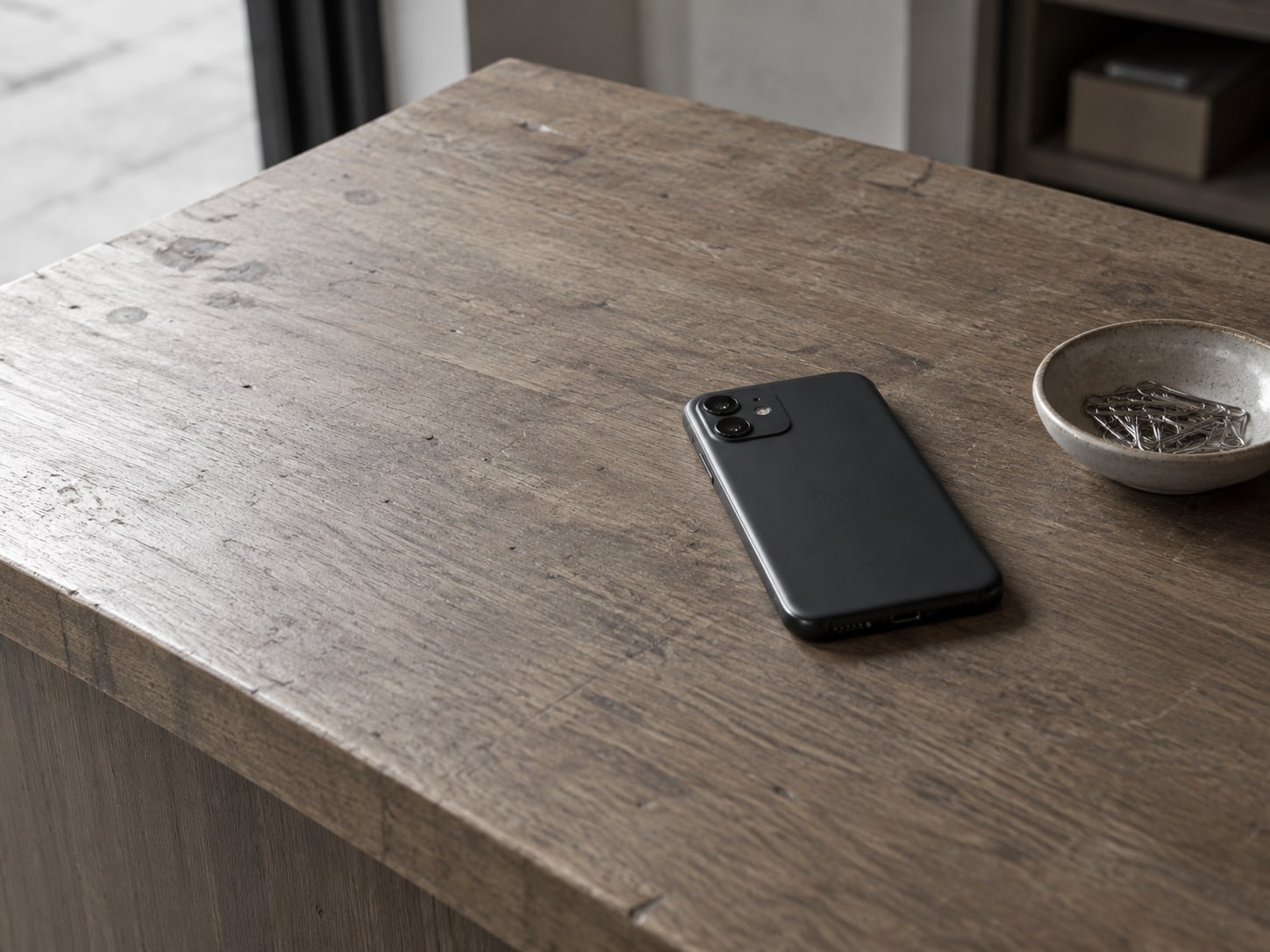 A single smartphone lying face down on a wooden shop counter beside a small ceramic dish of paper clips.