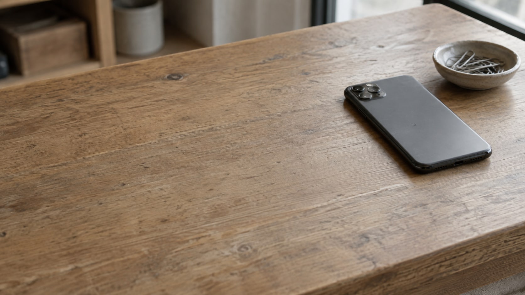 A smartphone lying face down on a wooden shop counter beside a small ceramic dish of paper clips.