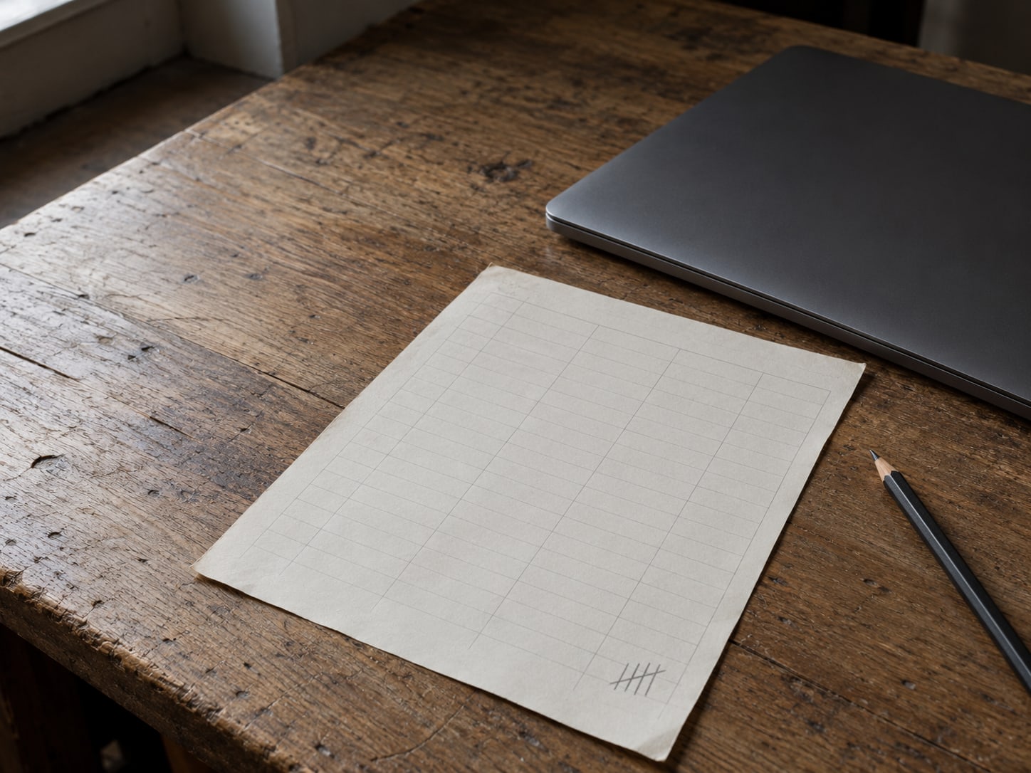 A printed sheet ruled in columns with pencil tally marks, set beside a closed laptop on a worn desk.