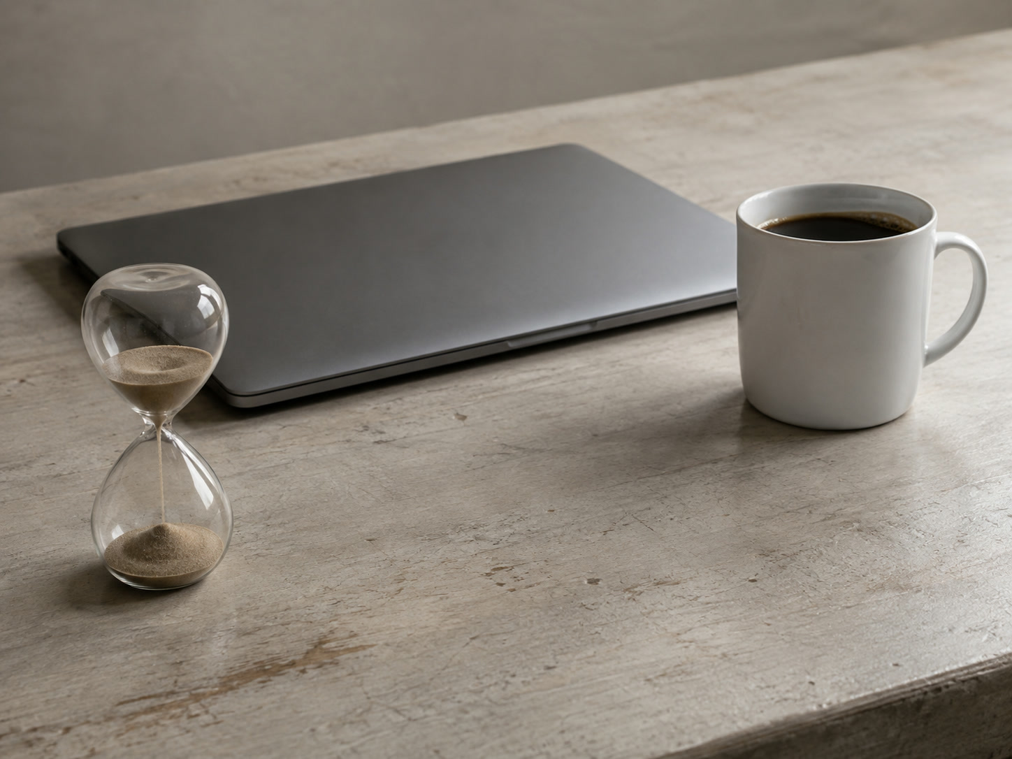 A small sand hourglass beside a closed laptop and a mug of coffee on a clean shop counter in soft morning light.