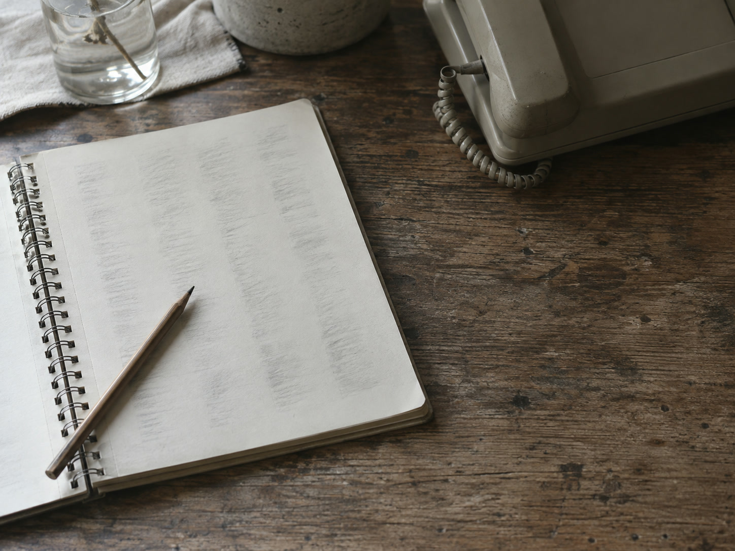 An open spiral notebook with faint, unreadable pencil columns, a pencil and a desk telephone on a wooden table.