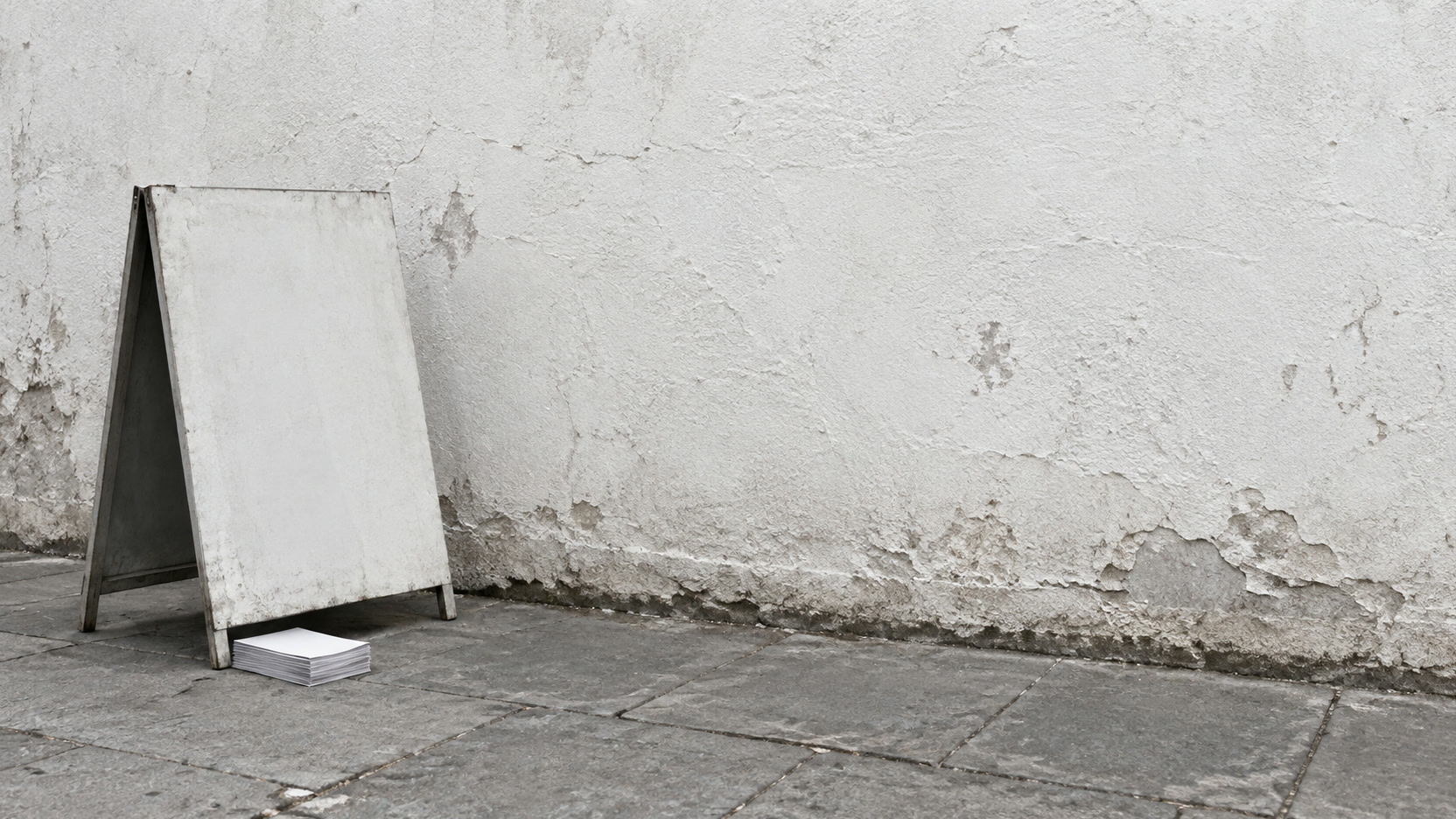 A blank sandwich-board sign folded against a shop wall beside a small stack of blank flyers.