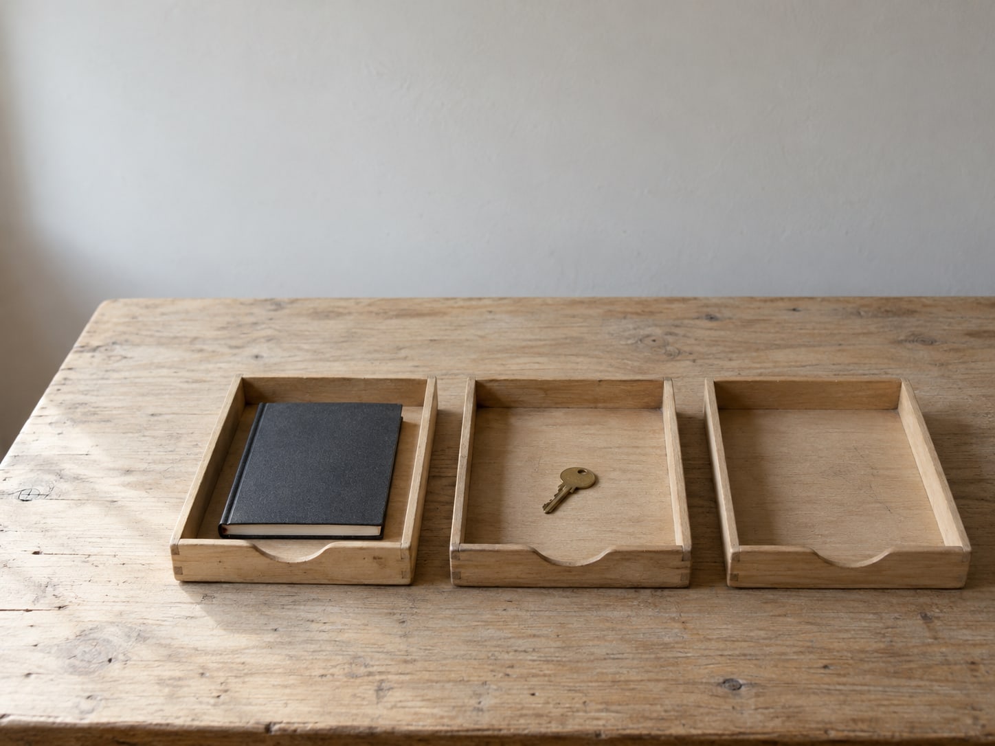 Tres bandejas de madera vacías sobre un escritorio: una con una libreta cerrada, otra con una pequeña llave de latón y la tercera vacía.