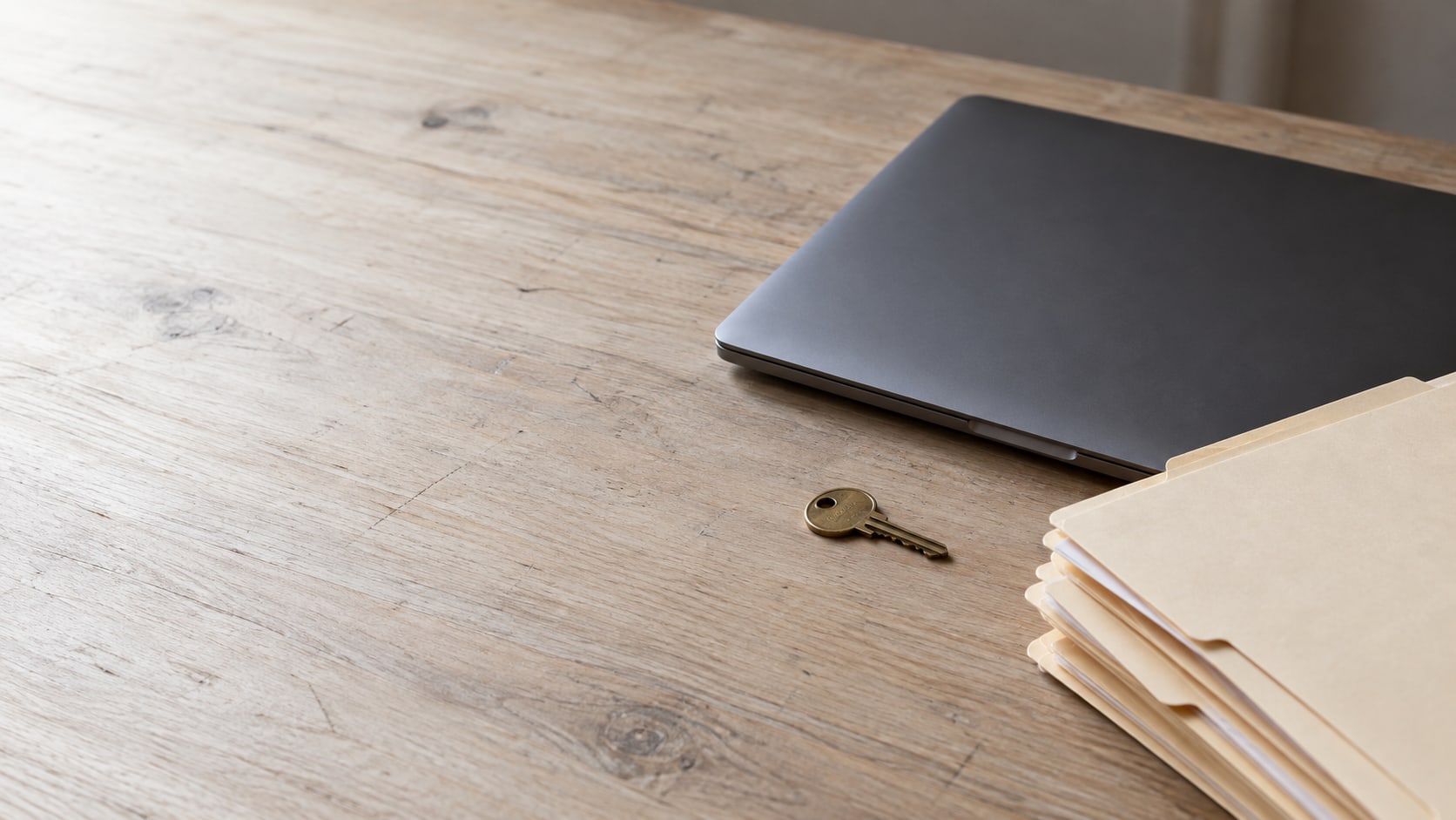 A closed laptop beside a small brass key and a stack of blank folders on a clean wooden desk.