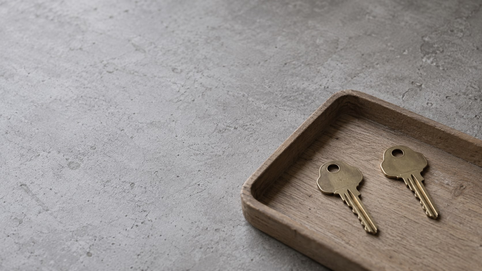 Two identical brass house keys resting on a small wooden tray, one placed slightly apart from the other.