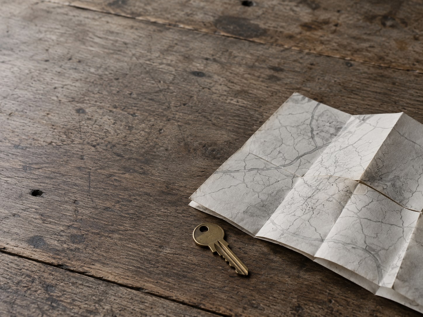 A folded paper map with faint pencil marks beside a brass key on a wooden table.