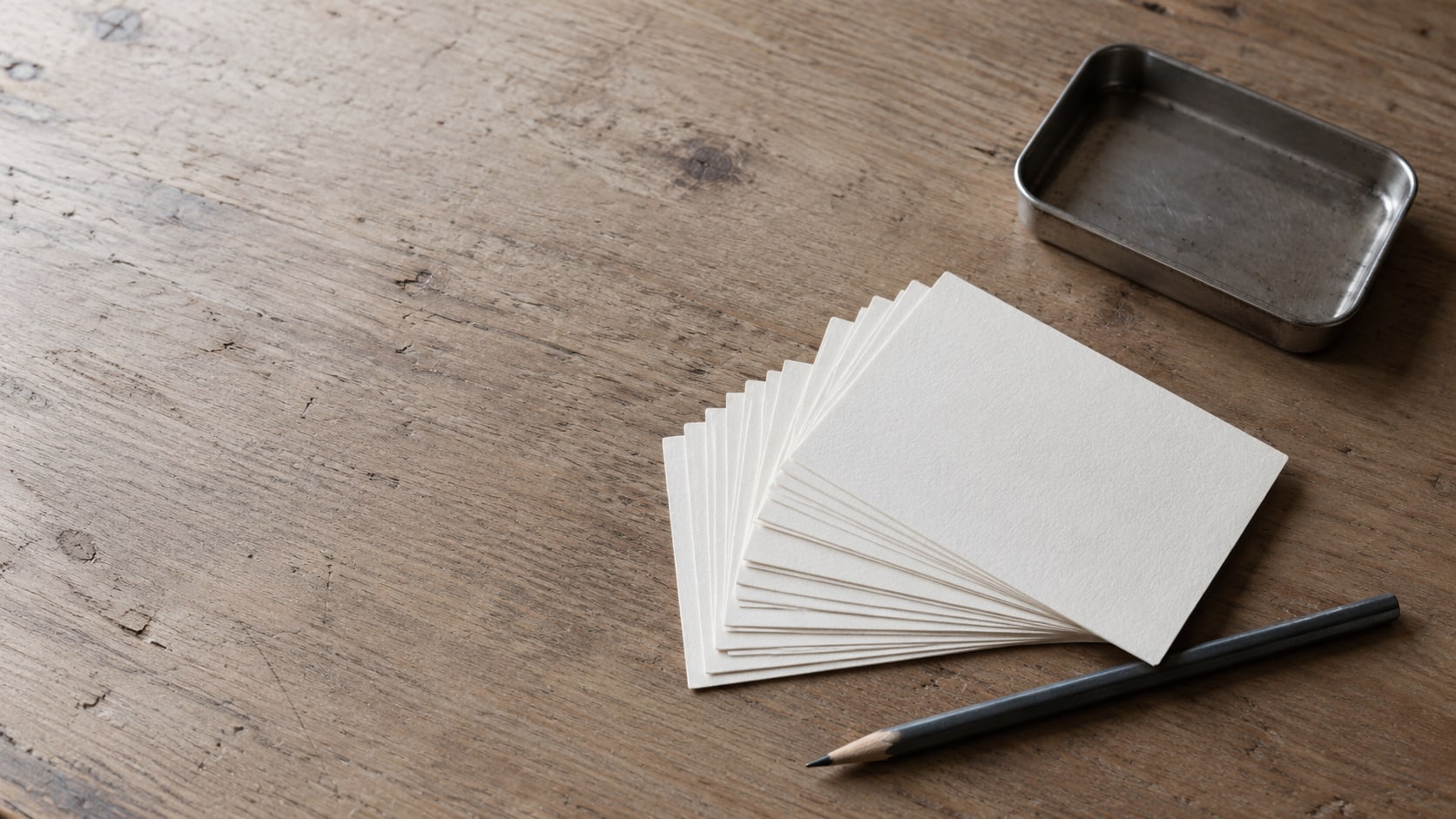 A fan of blank index cards beside a pencil and a small tray on a plain wooden desk.