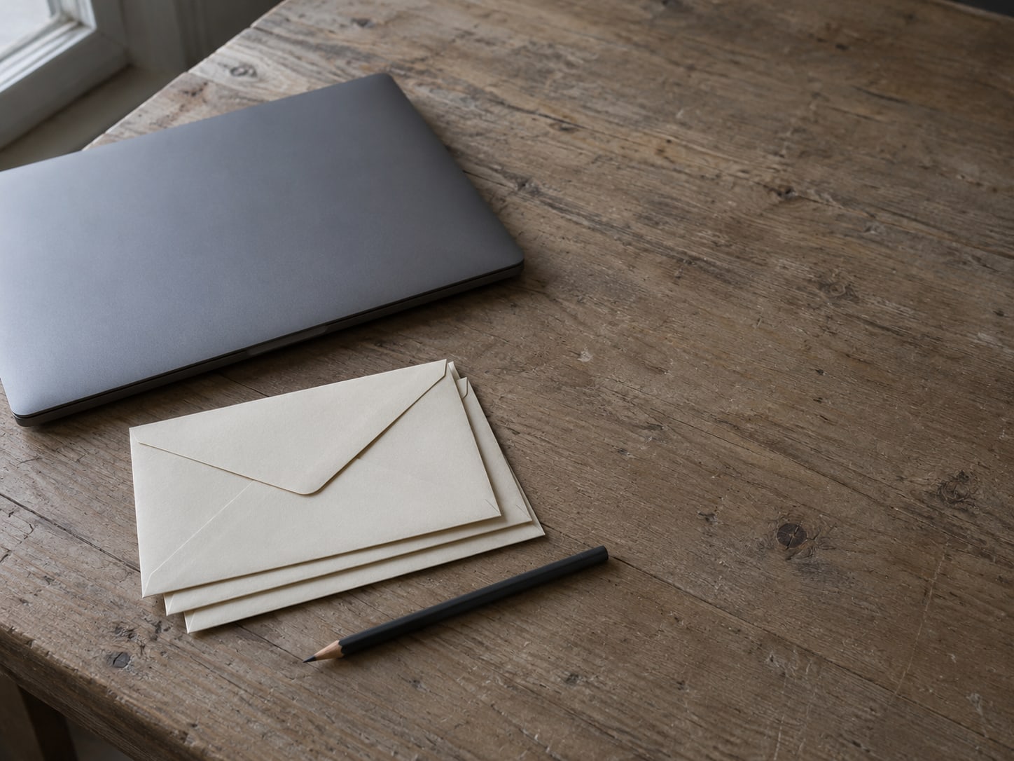 A closed laptop beside a short stack of unopened envelopes and a pencil on a worn wooden desk.
