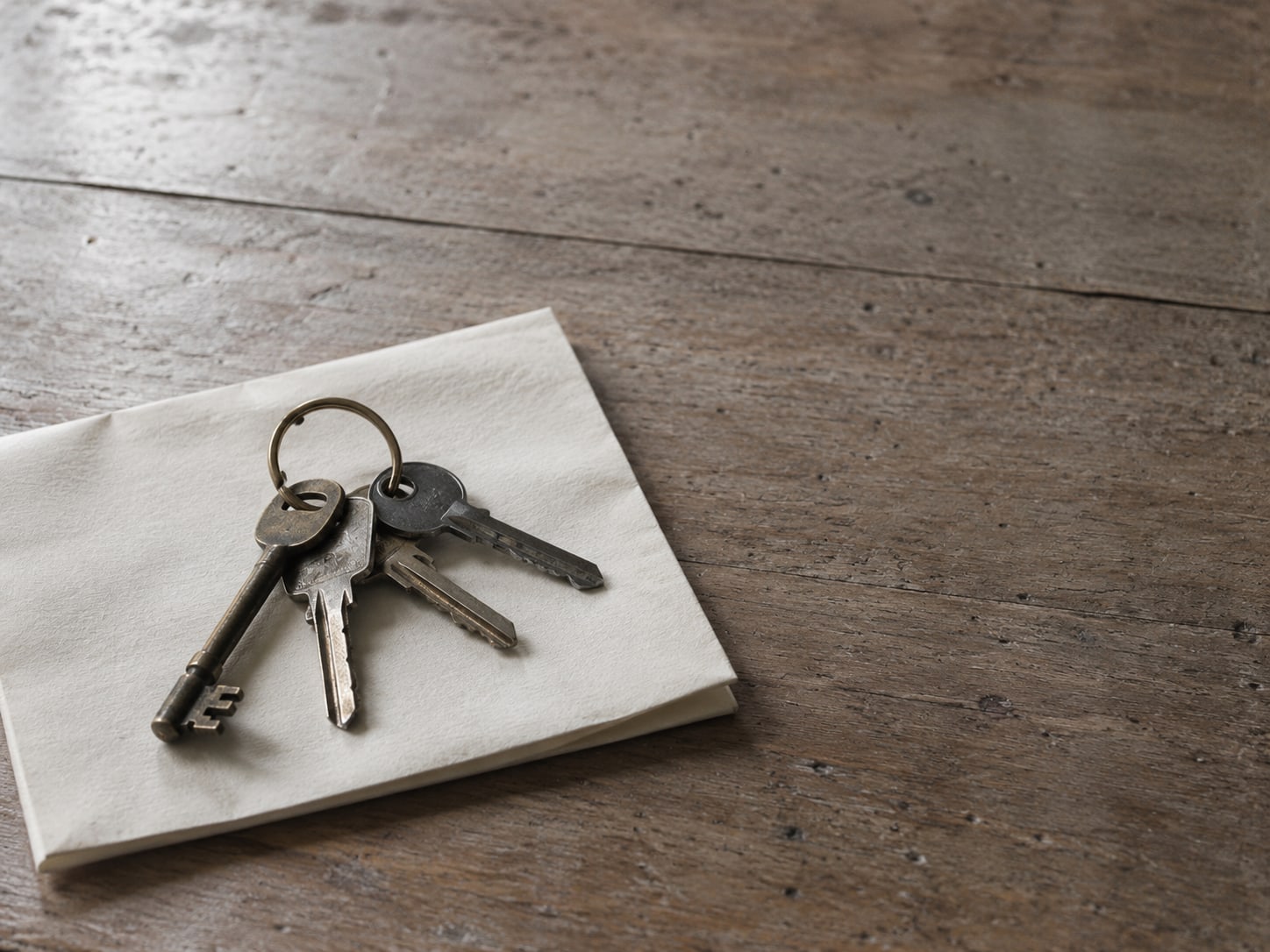 A small brass key ring holding four different keys, resting on a folded blank sheet of paper on a wooden desk.