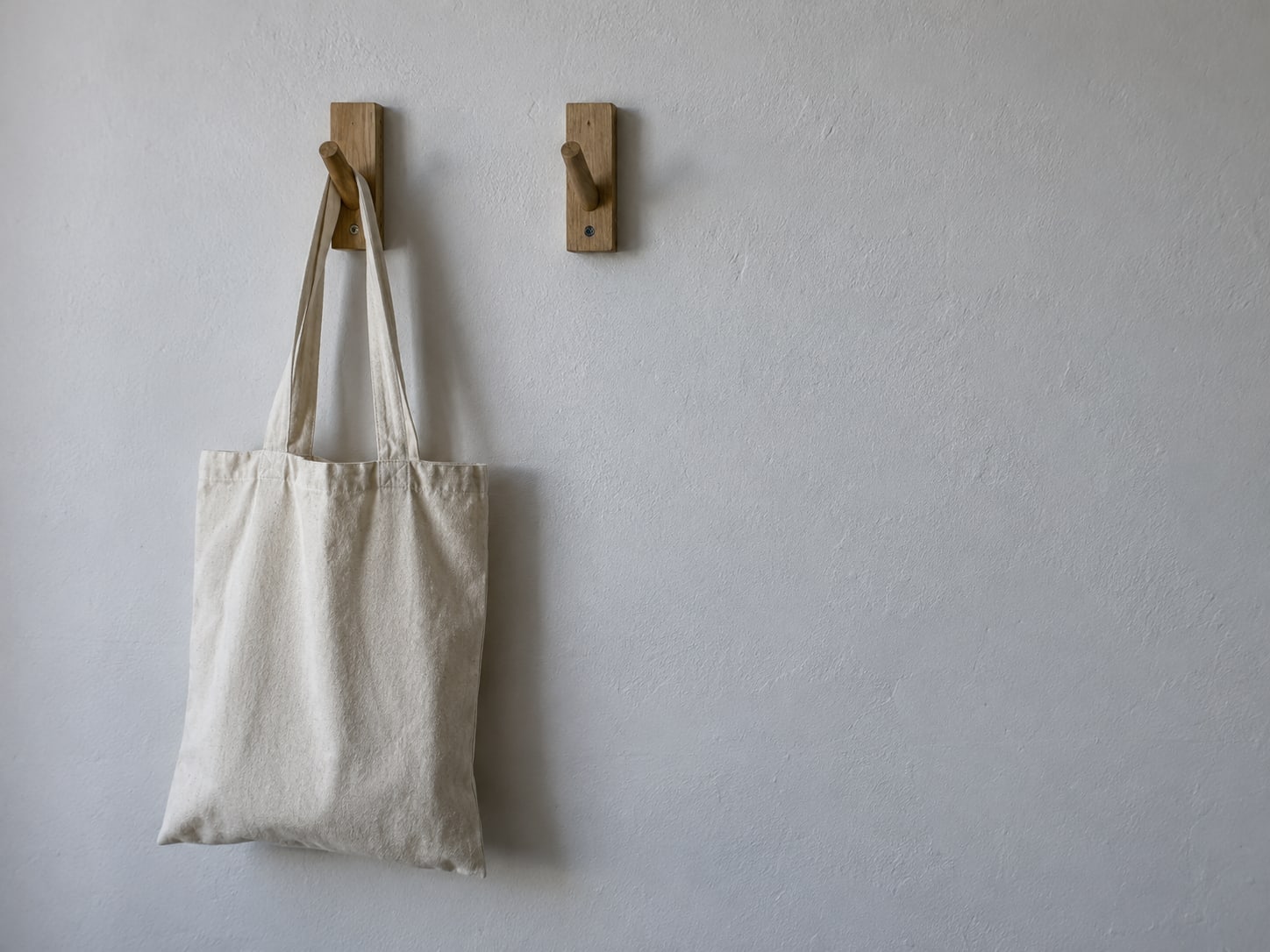 A pair of empty wooden coat hooks on a plain wall, one with a single canvas tote bag hanging from it.