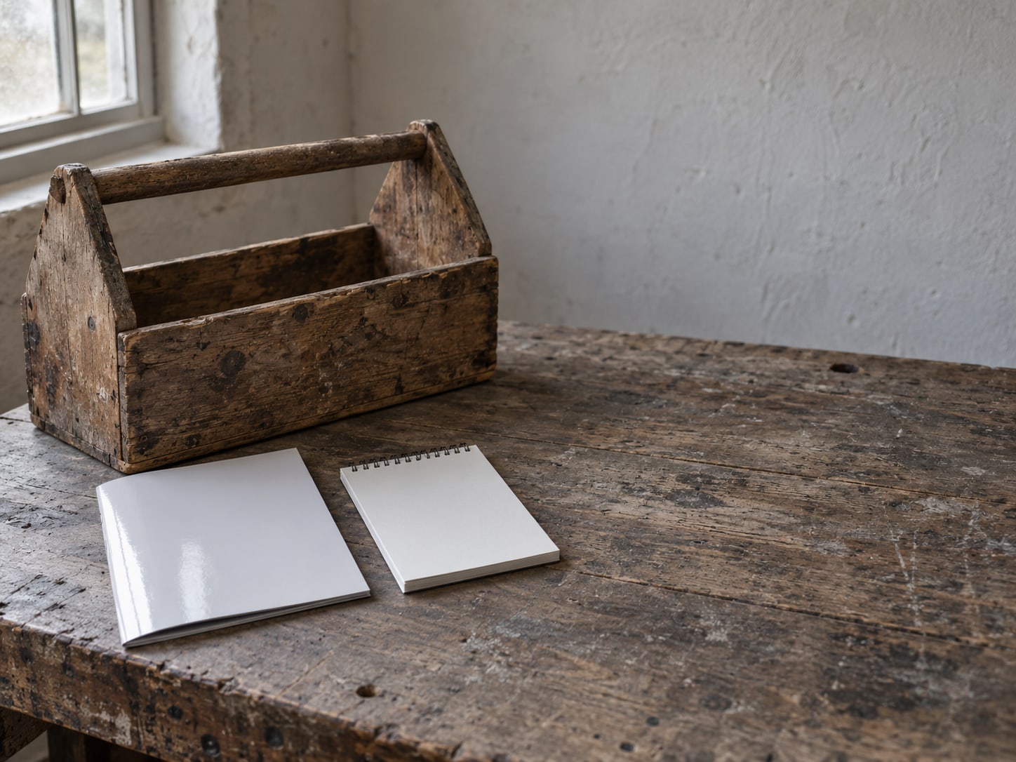 A battered wooden toolbox beside a glossy unopened brochure on a workbench, with a blank notepad between them.