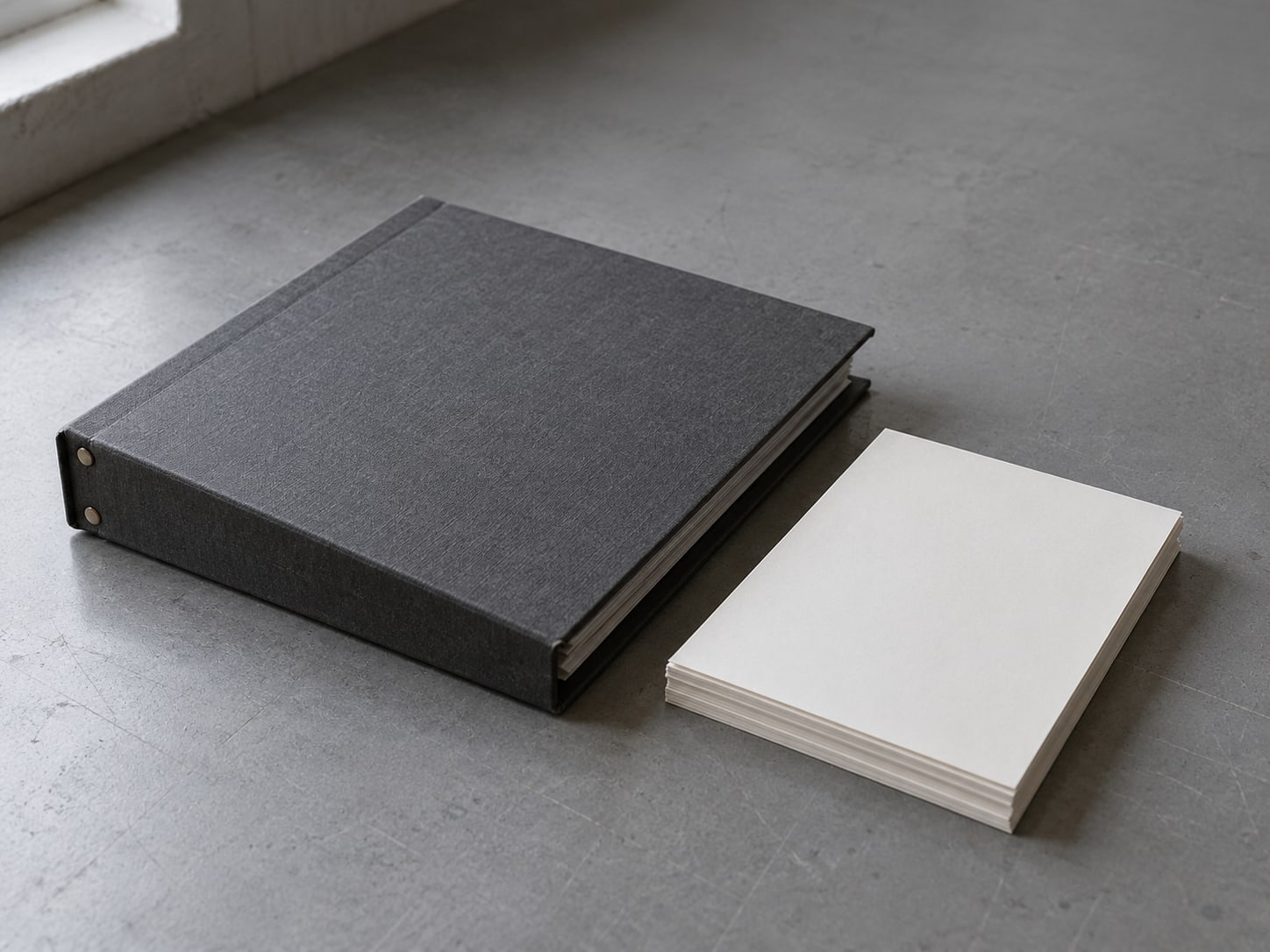 A tidy stack of identical blank paper sheets beside a single thick bound folder on a gray table.