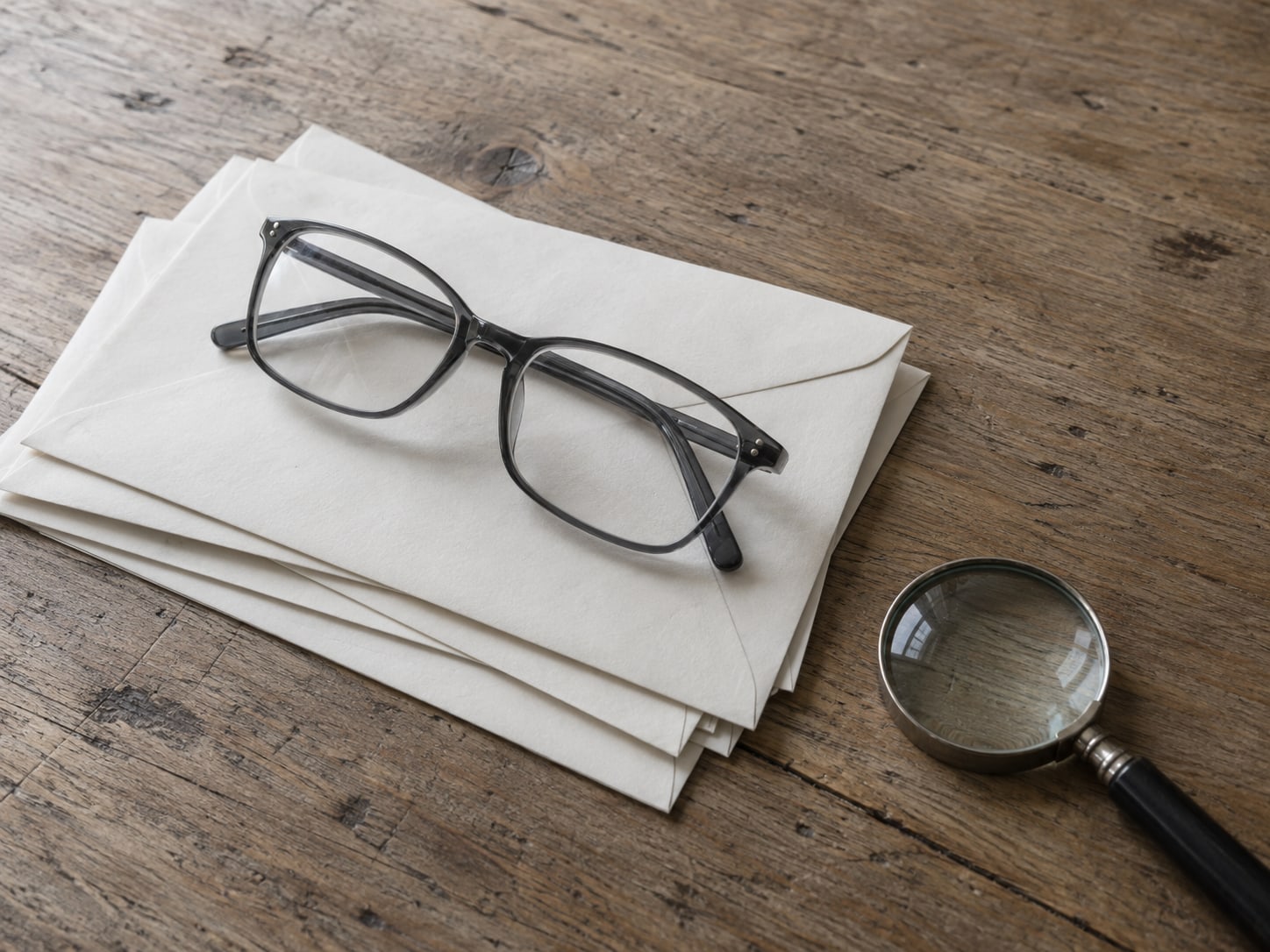 A pair of reading glasses lying on a stack of blank envelopes next to a small magnifying glass.