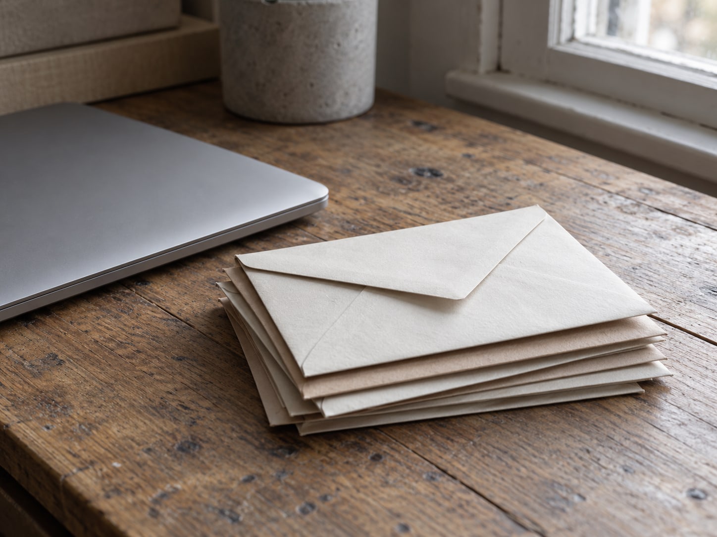 A closed laptop beside a small stack of unopened envelopes on a worn wooden desk.