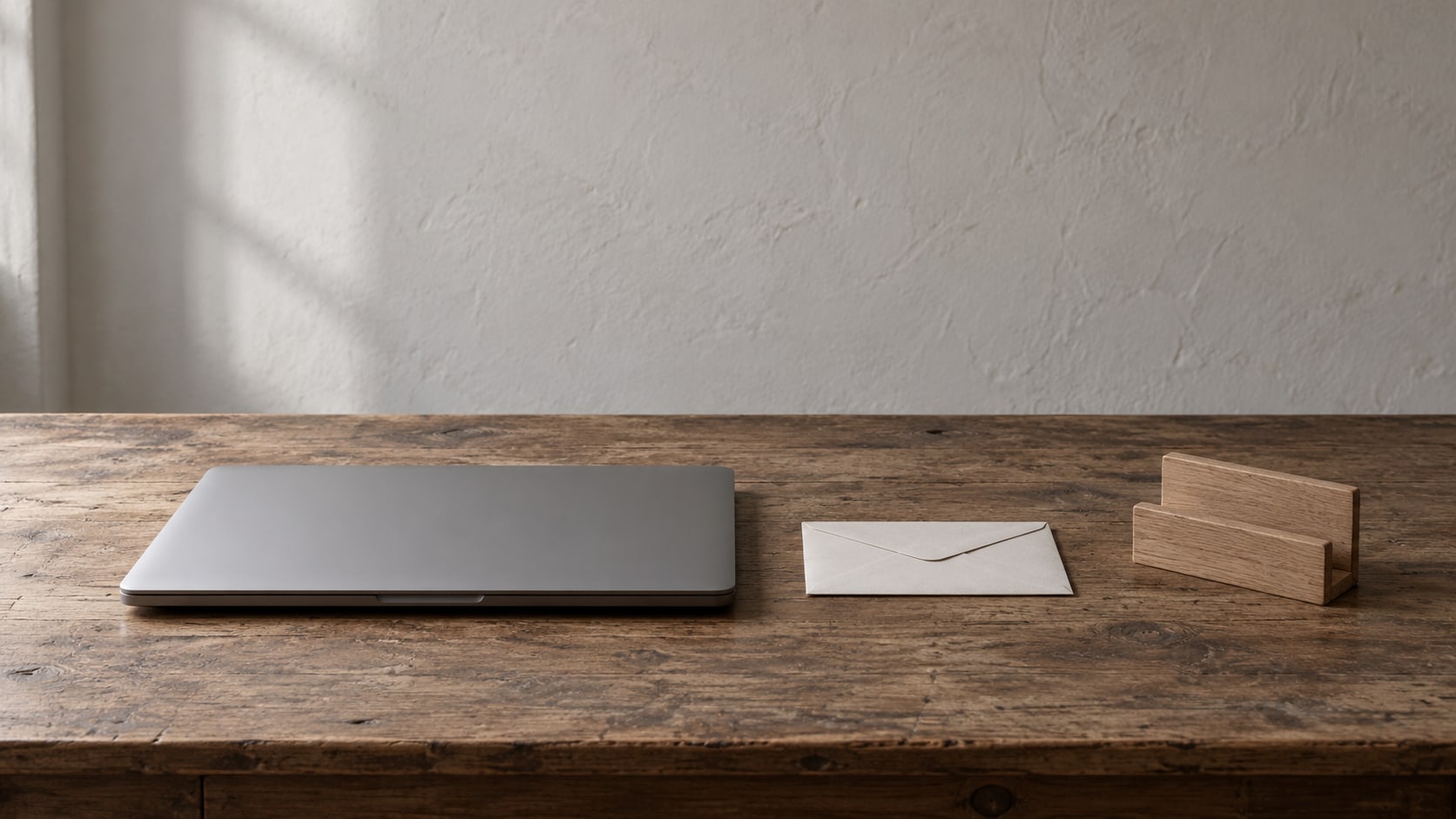 A closed laptop and a small sealed envelope beside a plain wooden card holder on a desk.