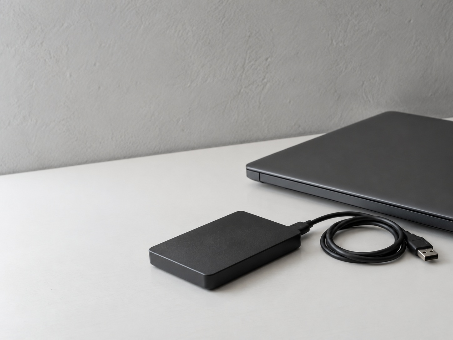 A small external hard drive with its cable coiled beside a closed laptop on a clean white desk.