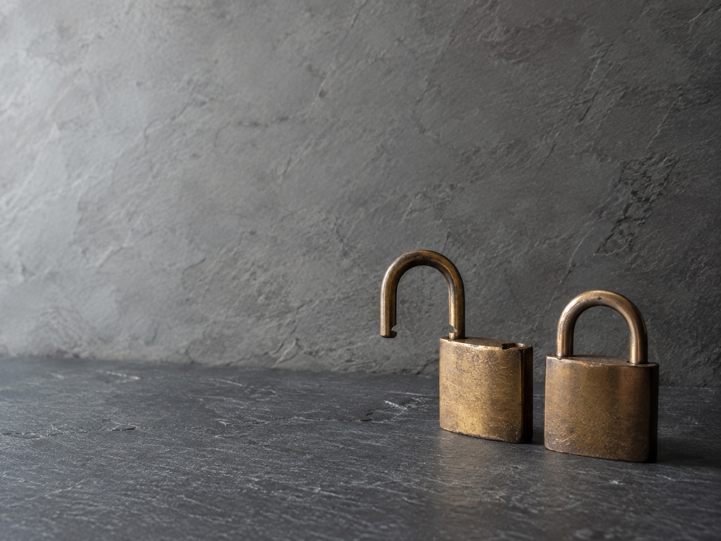 An open brass padlock resting beside a closed padlock on a dark slate surface.