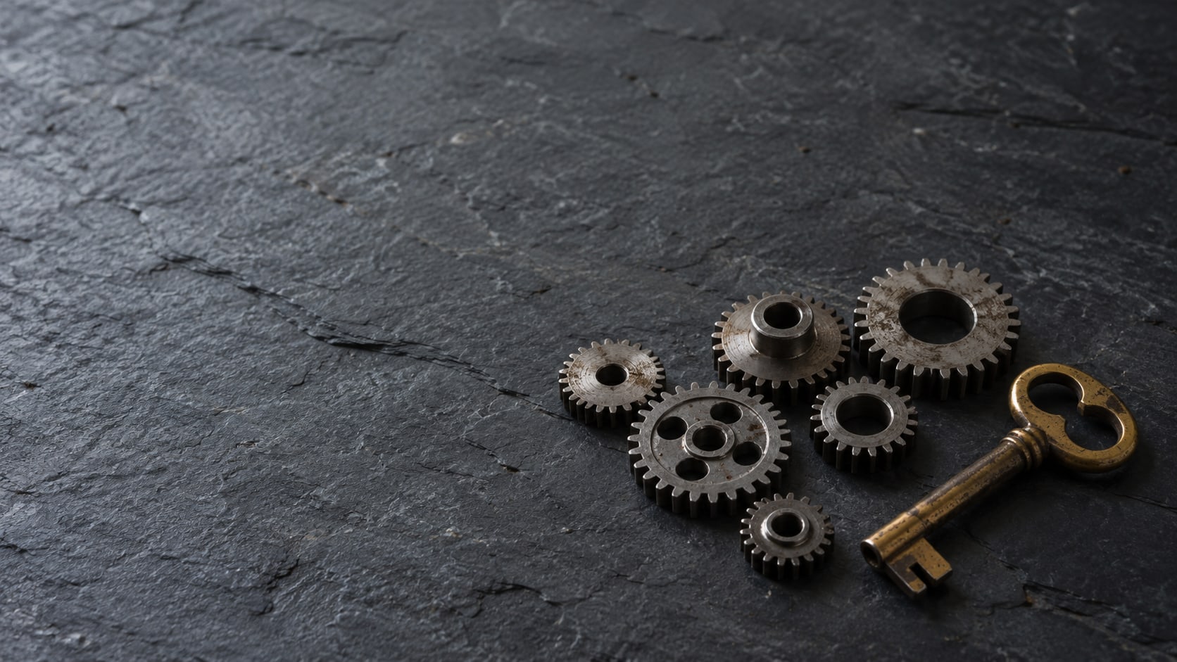 A row of small plain gears and a worn brass key laid out on a dark slate surface under soft light.