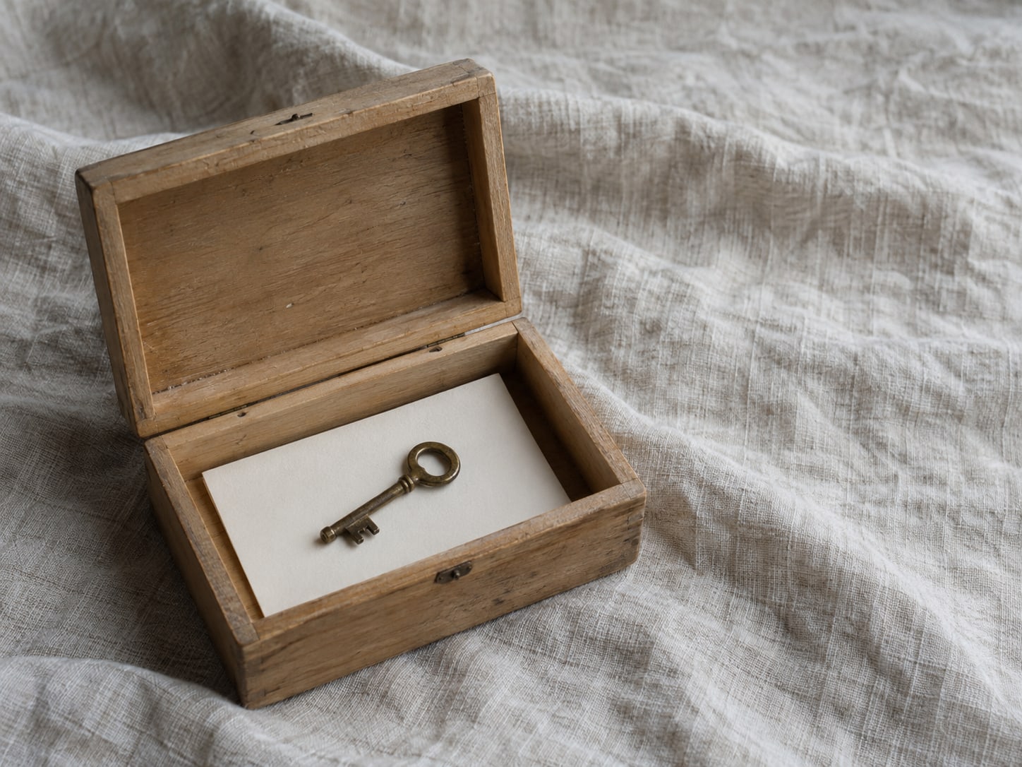 A small brass key on top of a blank index card inside an open wooden recipe box on a linen cloth.