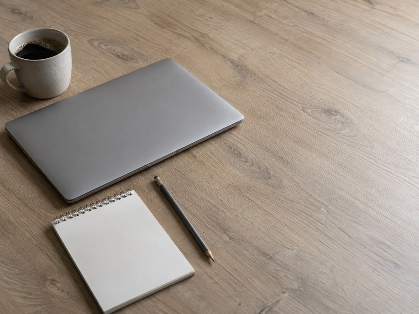 A closed laptop beside a small notepad with a pencil and a cup of coffee on a plain oak desk.