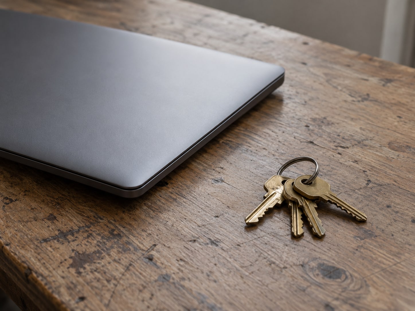 A closed laptop beside a ring of unused brass keys on a worn wooden desk.