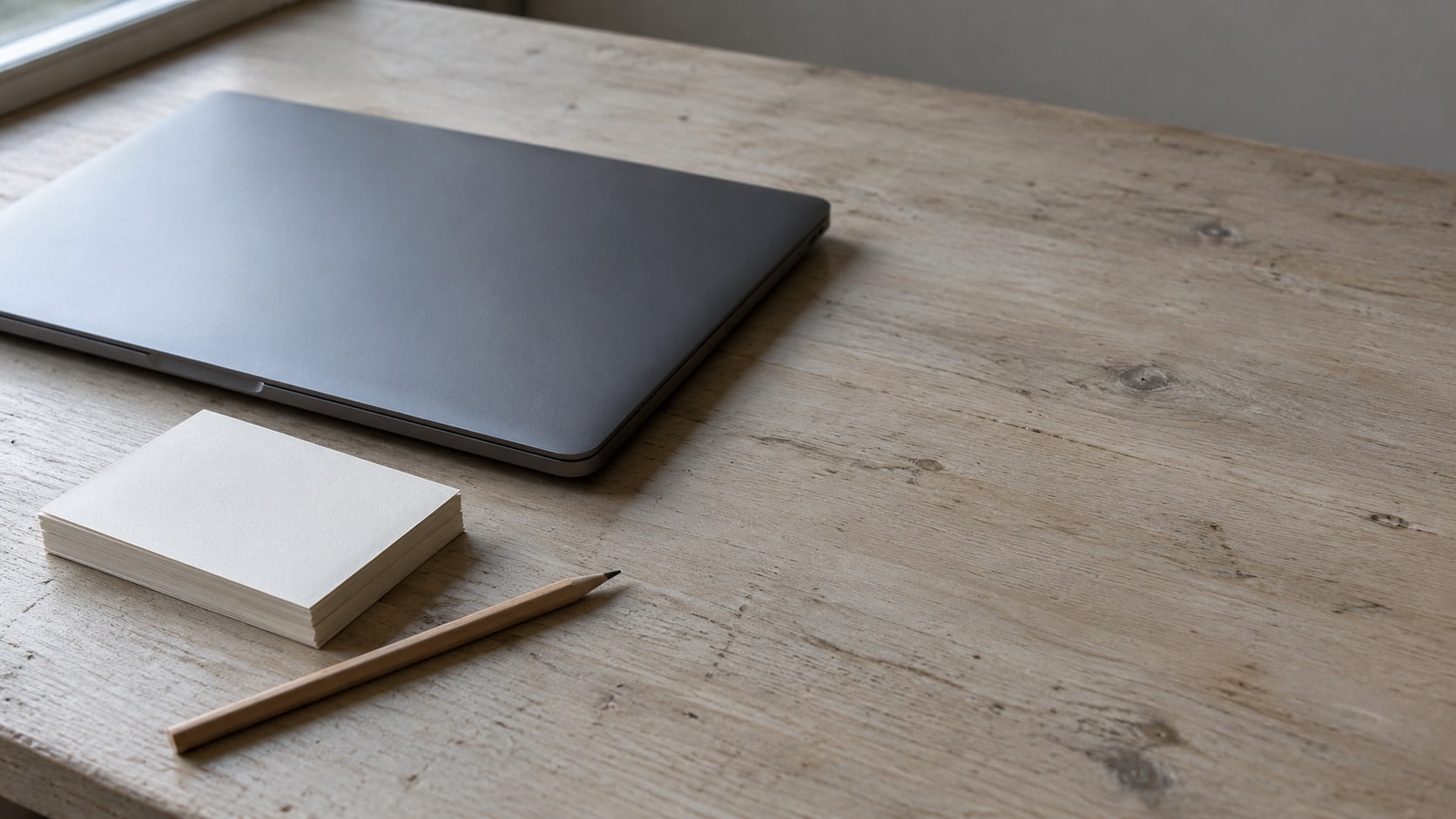 Una laptop cerrada, una pila de tarjetas de índice en blanco y un lápiz dispuestos sobre un escritorio de madera clara.