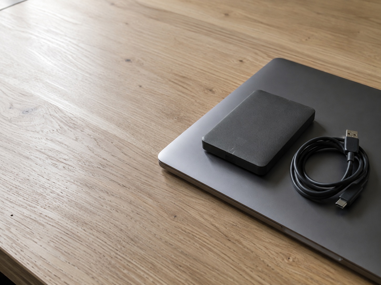 An external hard drive resting on top of a closed laptop on a plain oak desk, with a coiled cable beside it.